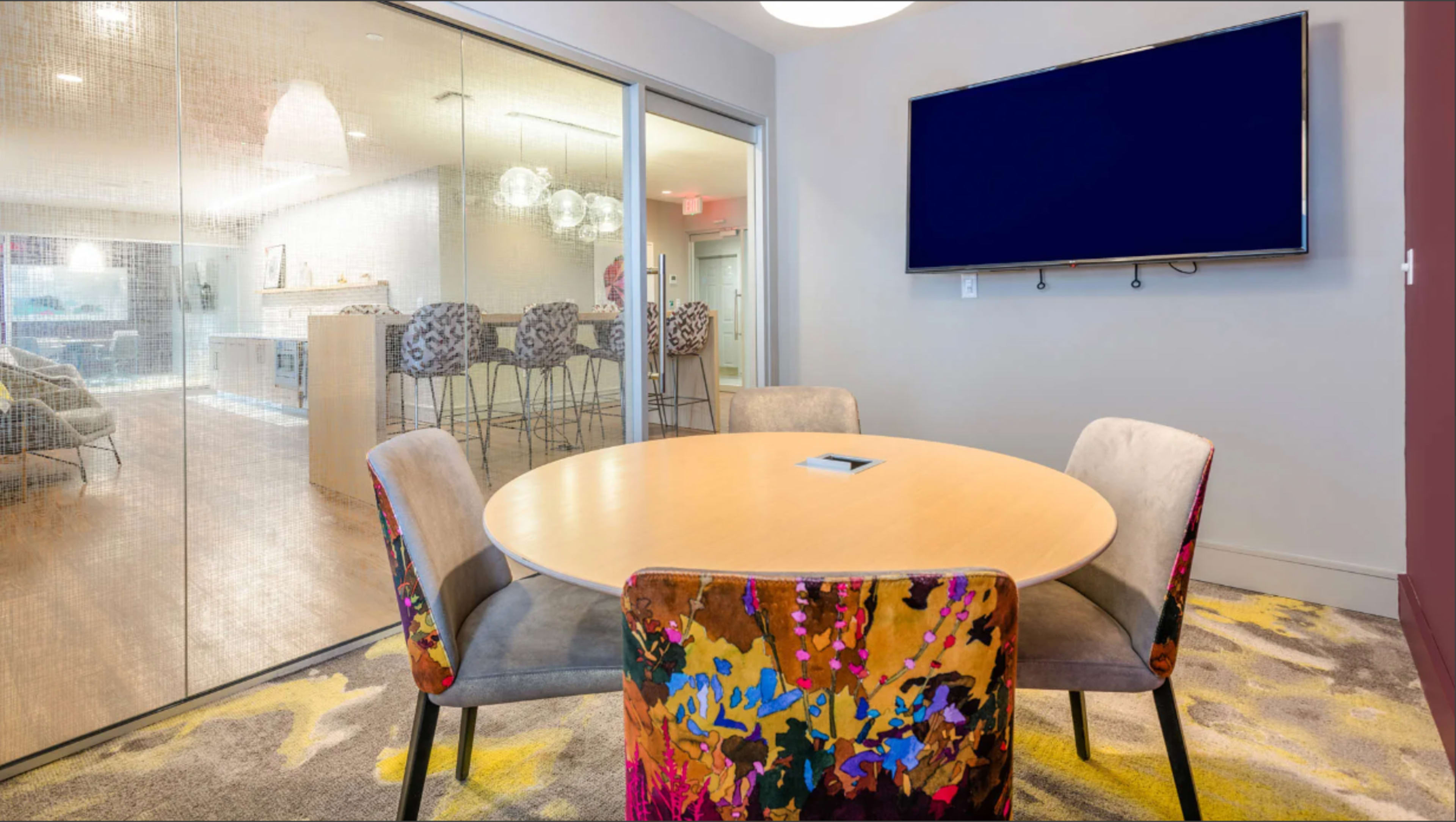 A round table with colorful chairs is set in a modern meeting room, adjacent to a space featuring a kitchen and bar stools, with a large screen mounted on the wall.