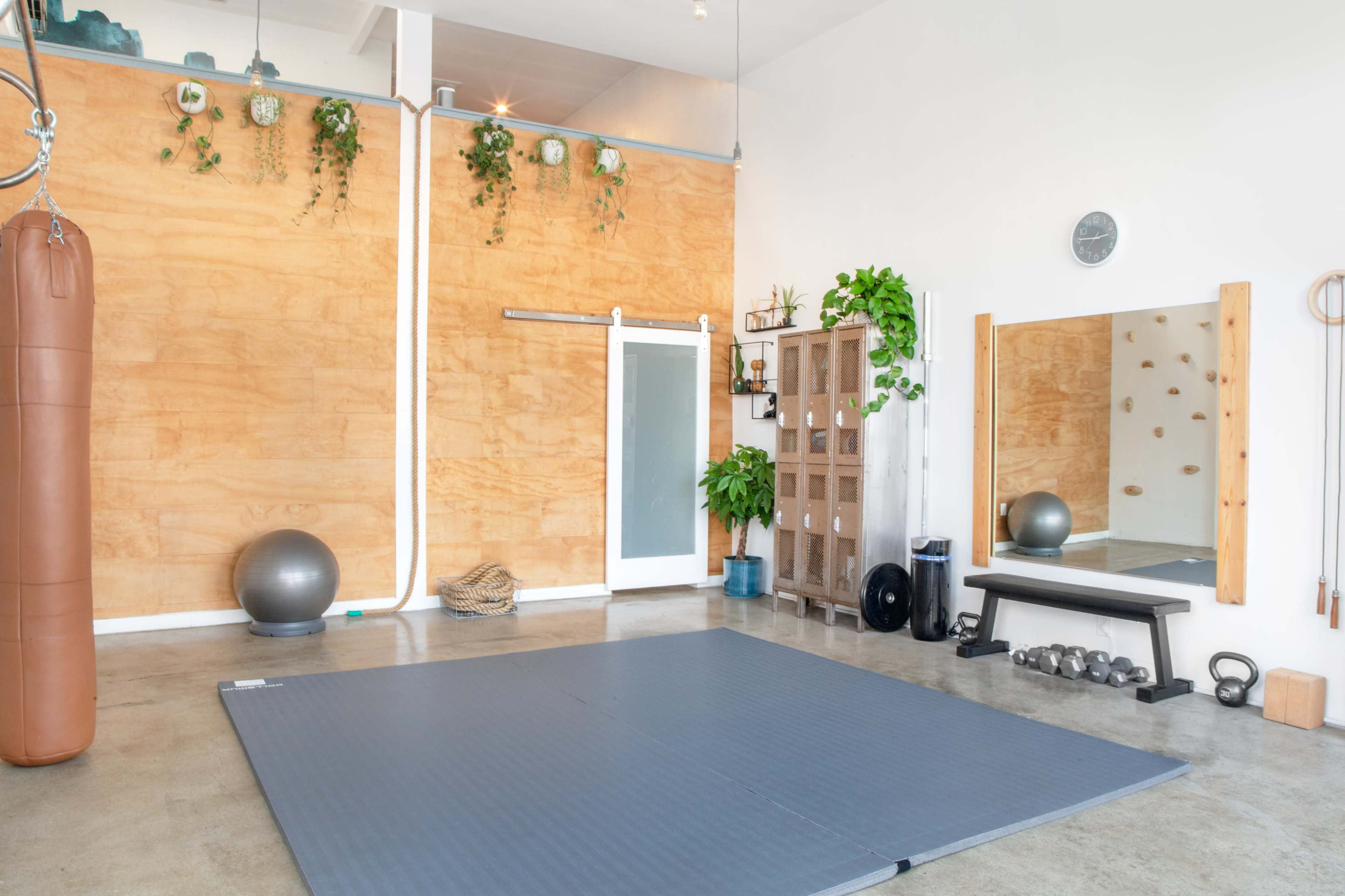 A spacious, well-lit workout area with a blue exercise mat, a punching bag, a mirror, and various fitness equipment against a wooden wall.