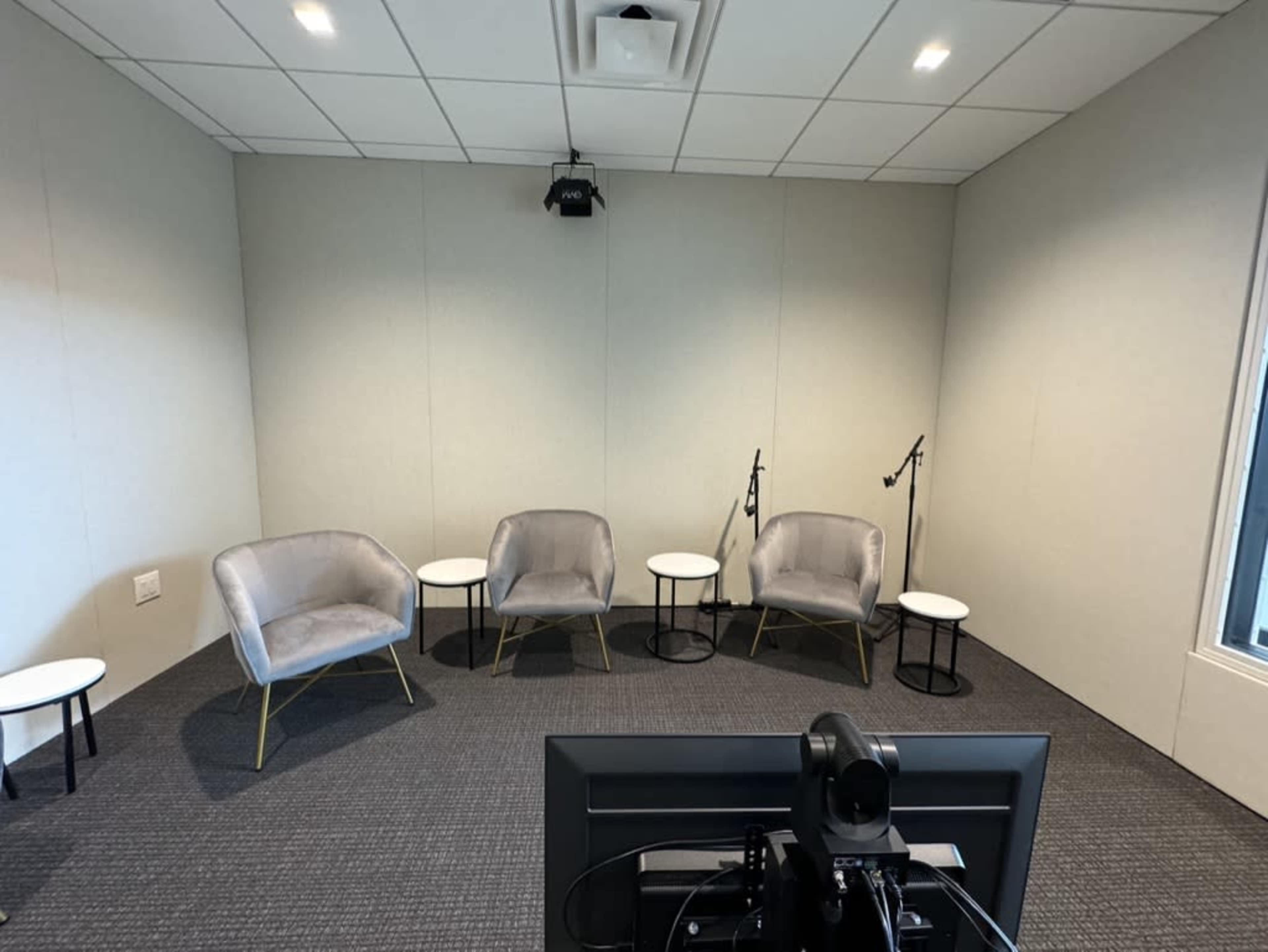 The image shows a small, minimally furnished room with four chairs and two small tables arranged around a central area, facing a monitor.