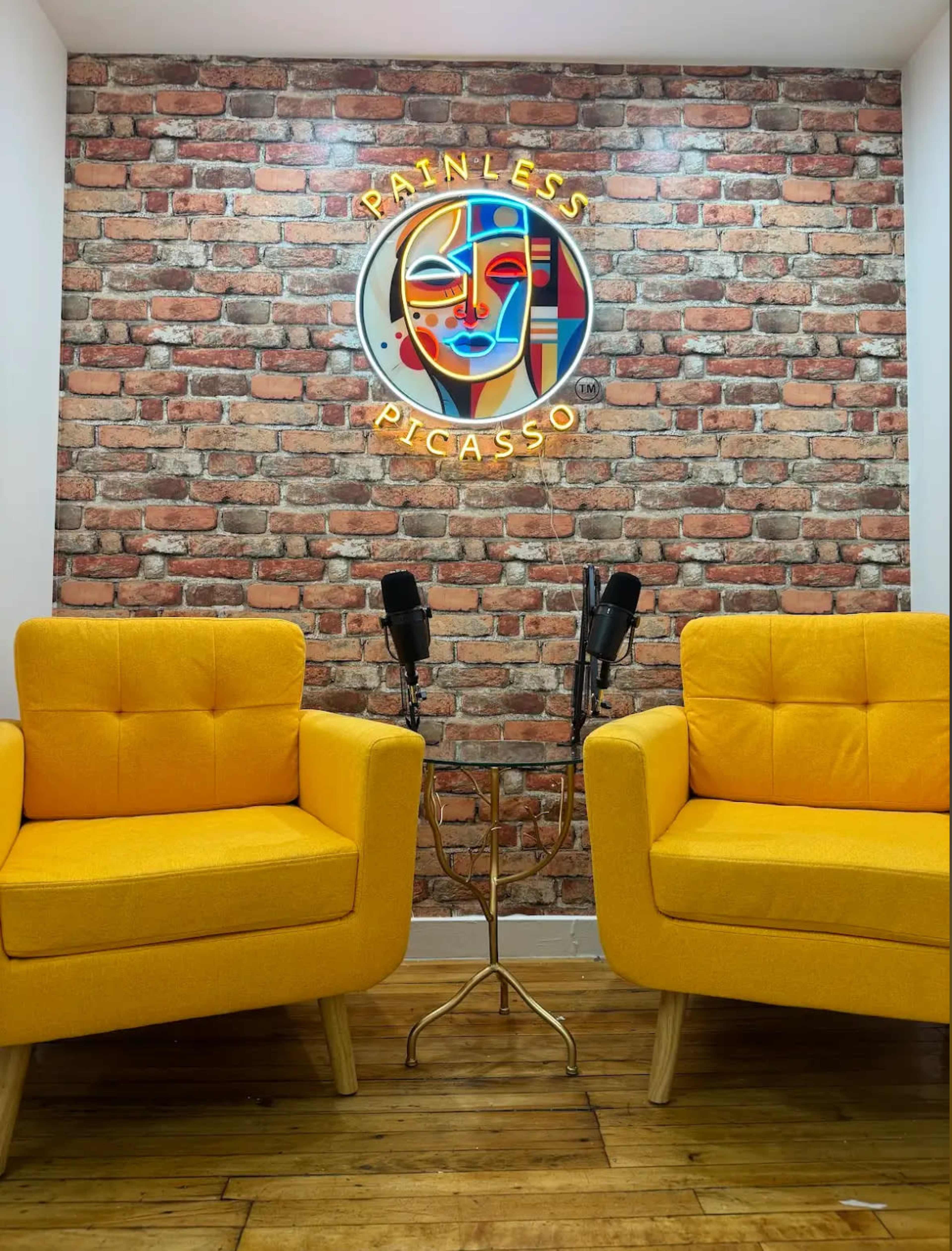 The image shows two yellow chairs facing each other in front of a brick wall with a neon sign that reads "Painless Picasso."