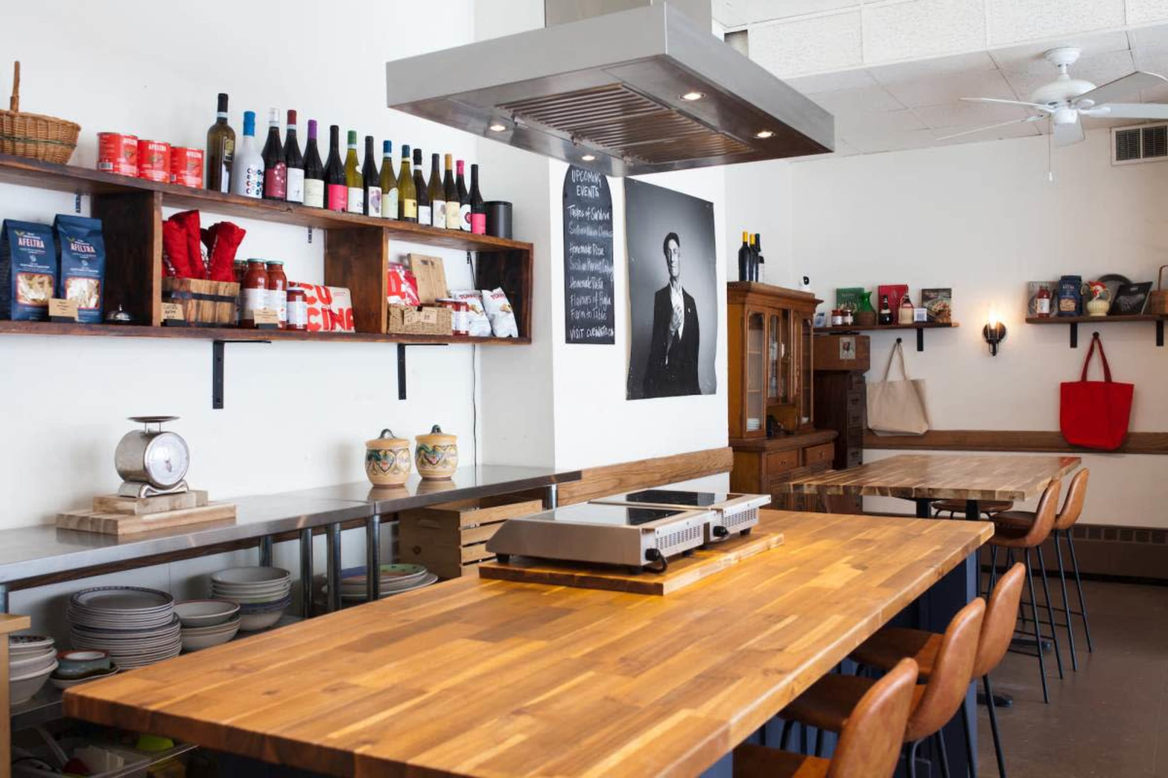 The image shows a modern kitchen space with wooden tables, shelves filled with various cooking ingredients, and a large poster on the wall.