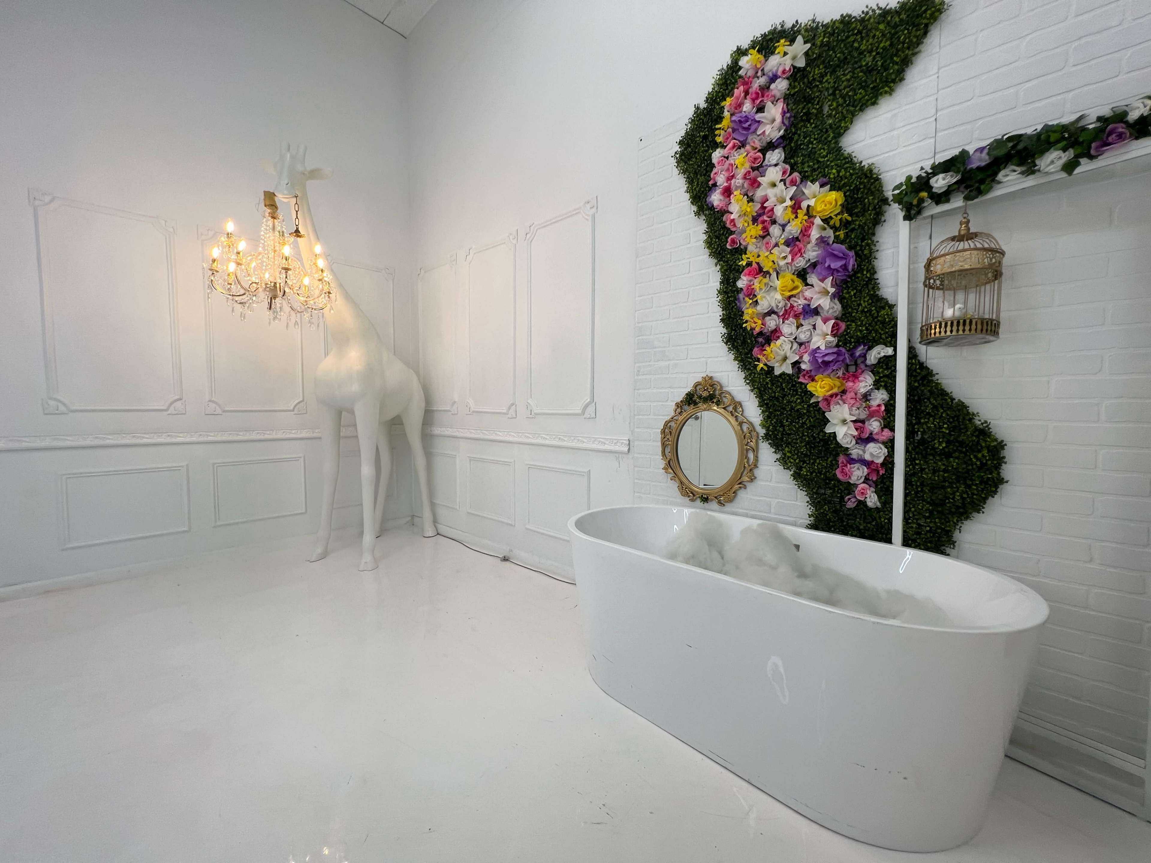 A modern bathroom with a freestanding white bathtub, a decorative wall of flowers and greenery, a large white giraffe sculpture, and a chandelier.