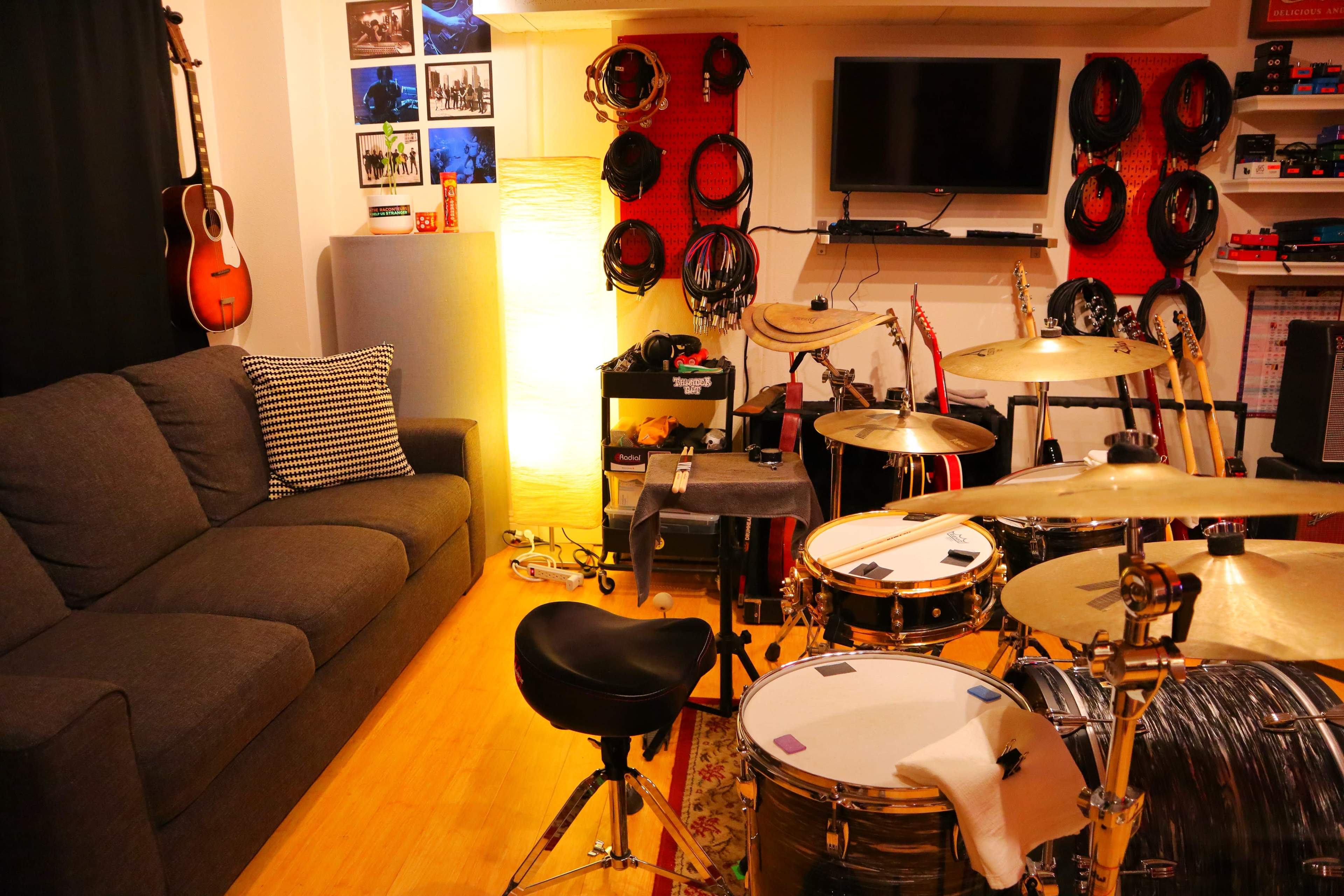 The image shows a music rehearsal space featuring a cozy couch, a drum set, and various instruments displayed on the walls.