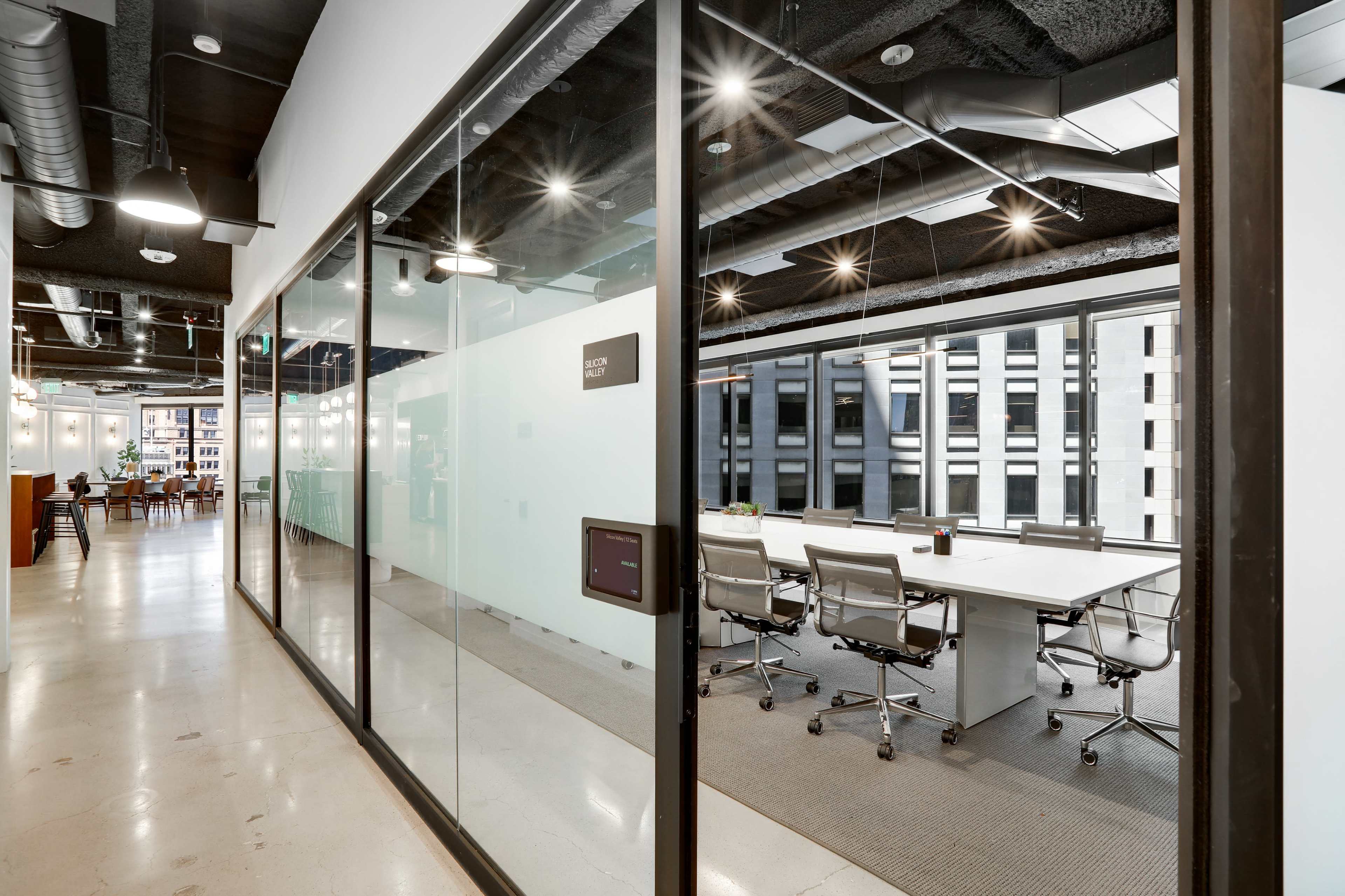 A modern office space featuring a glass-walled conference room with a large table and chairs, alongside an open hallway with additional work areas.