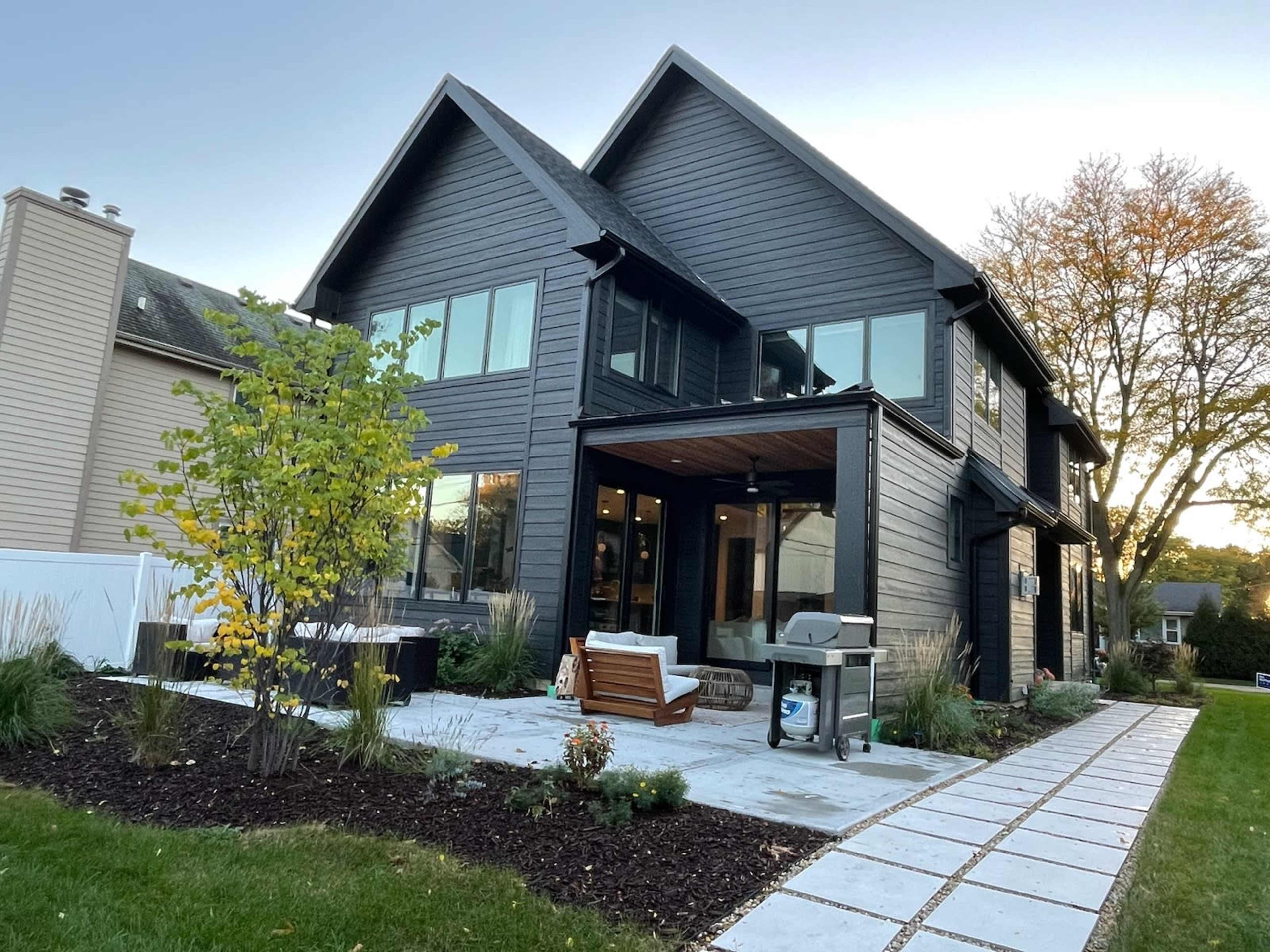 A modern black wooden house with large windows, a covered porch, and a neatly landscaped yard featuring a pathway and plants.