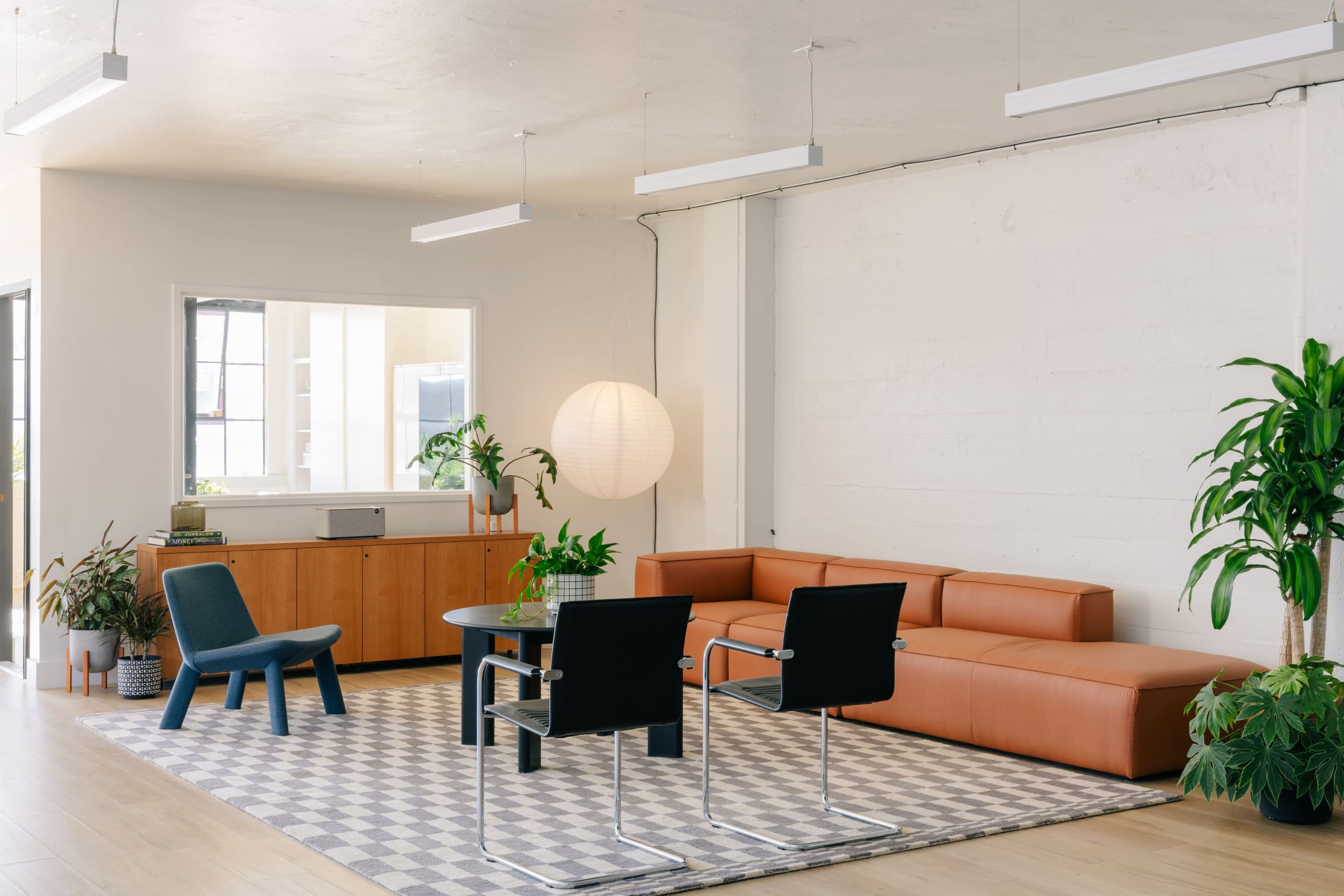 The image shows a modern office interior featuring a light-colored floor, a brown sectional sofa, a round table with four black chairs, and decorative plants.