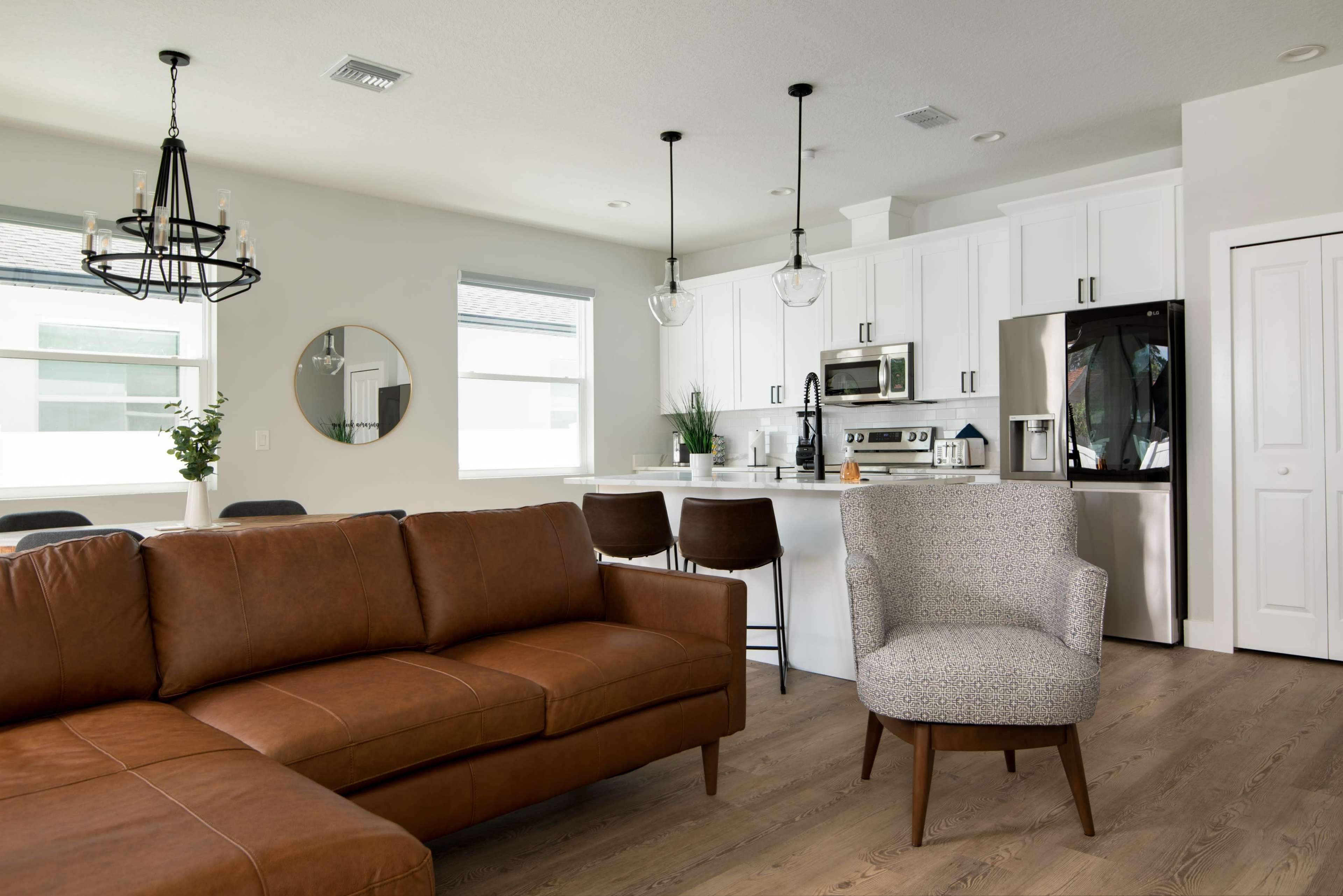 A modern kitchen and living area featuring a brown leather sofa, a round dining table, and white cabinetry with stainless steel appliances.