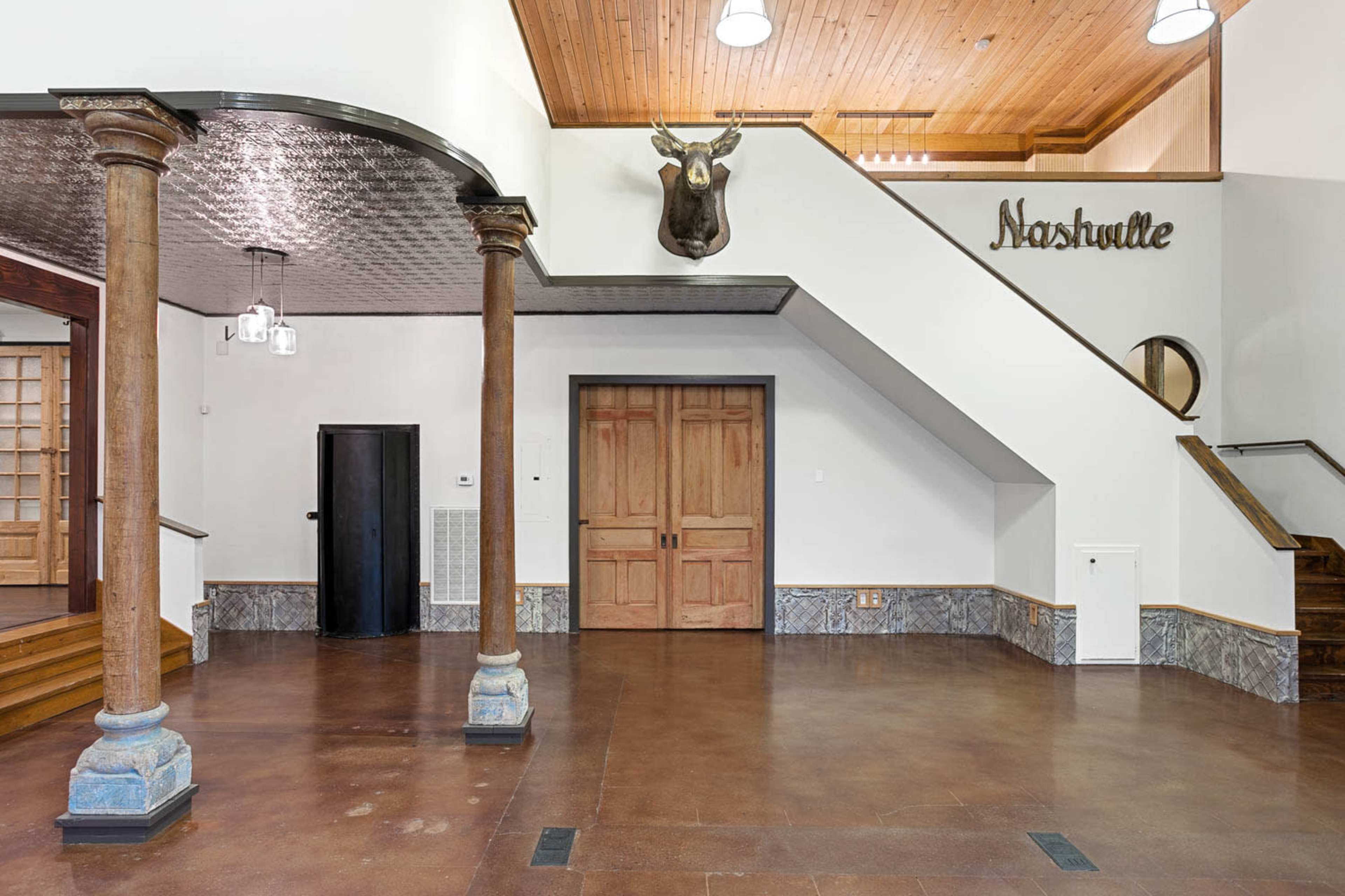 The image shows a well-lit entryway featuring wooden columns, a mounted moose head, and a staircase leading to an upper level.
