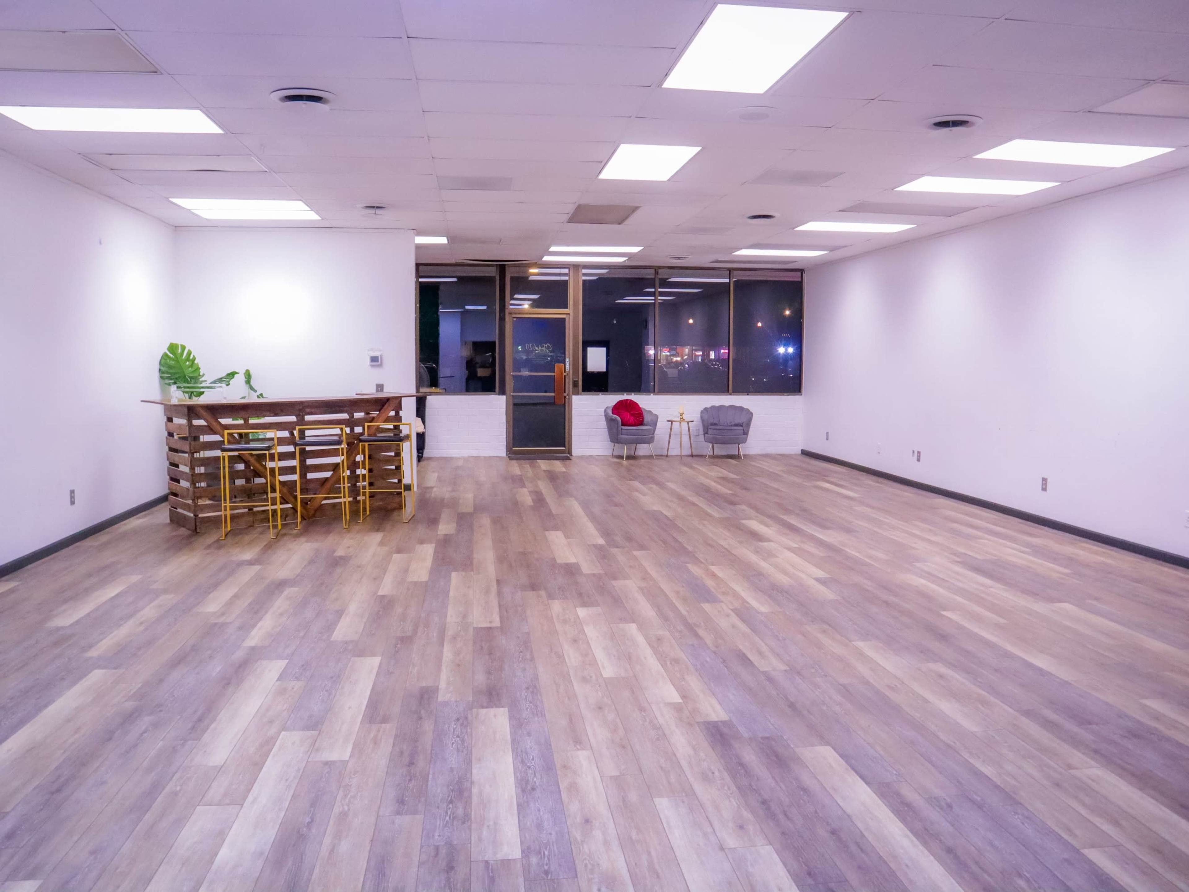 An empty room with wooden flooring, featuring a small dining area with yellow chairs and a wooden table on one side, and a seating area with a gray armchair on the opposite side.
