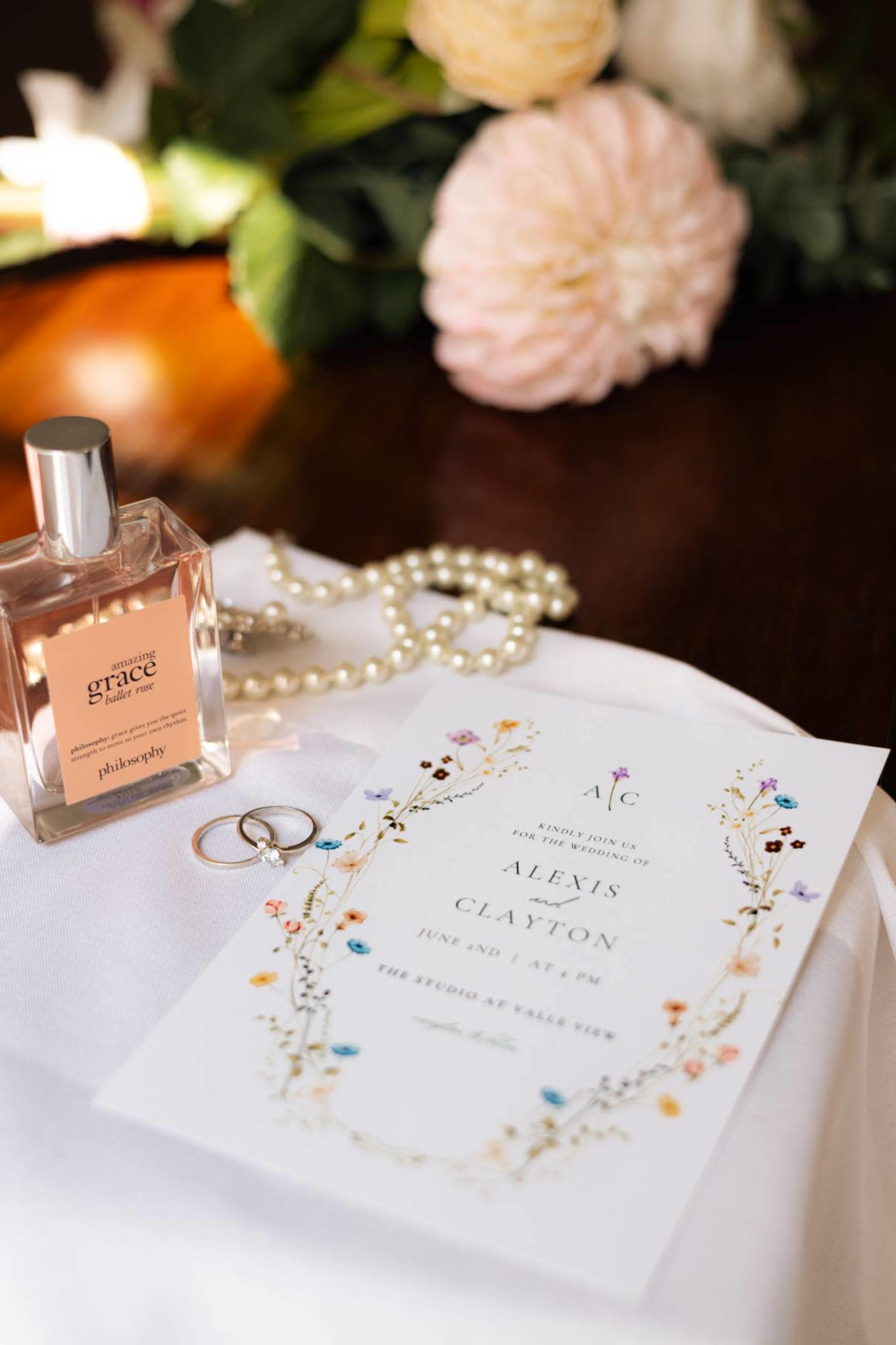 The image features a wedding invitation surrounded by a perfume bottle, a string of pearls, and two rings on a white cloth.