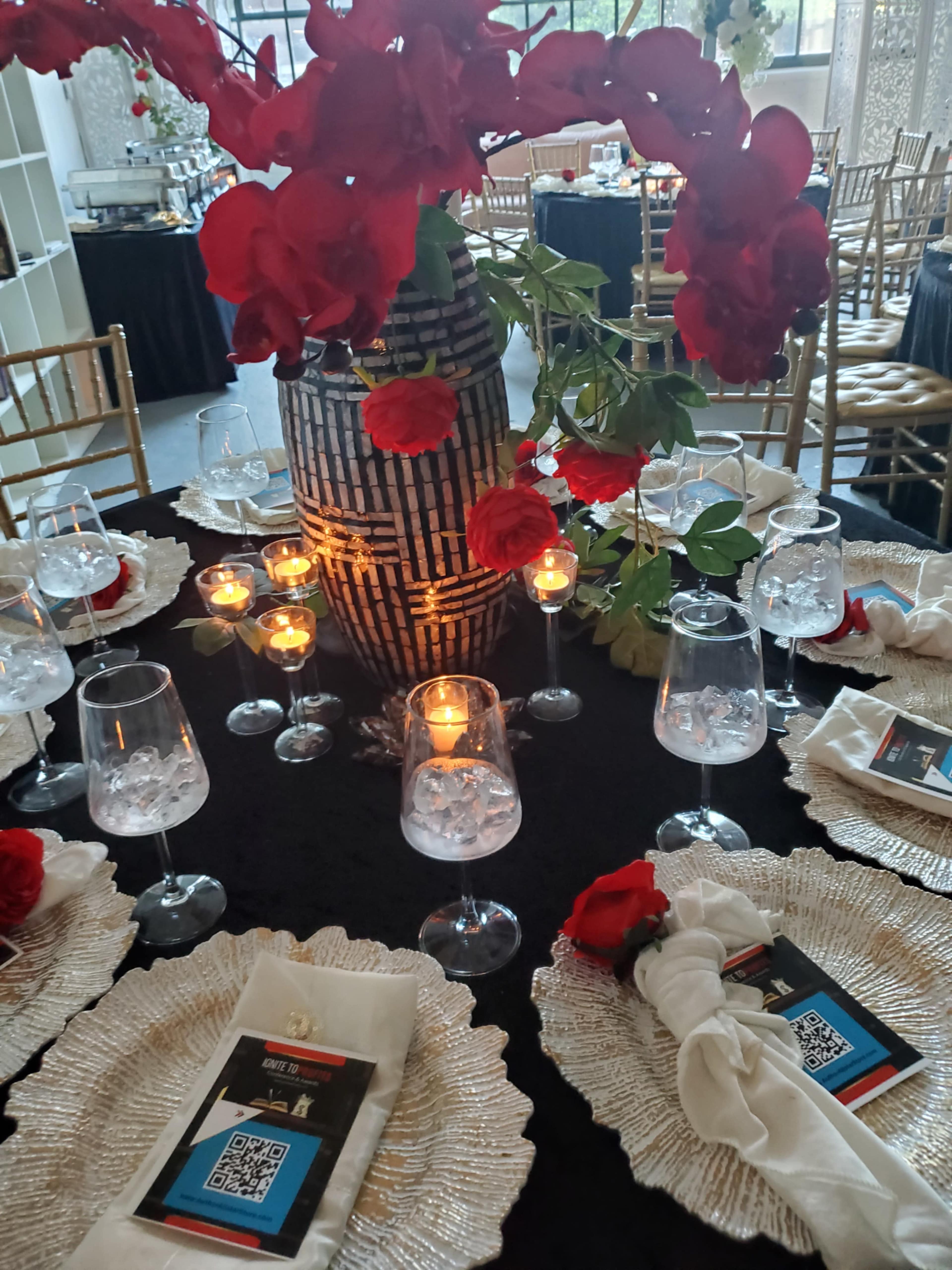 A table is elegantly set with white placemats, wine glasses, and menus, centered around a decorative vase filled with red flowers and illuminated candles.