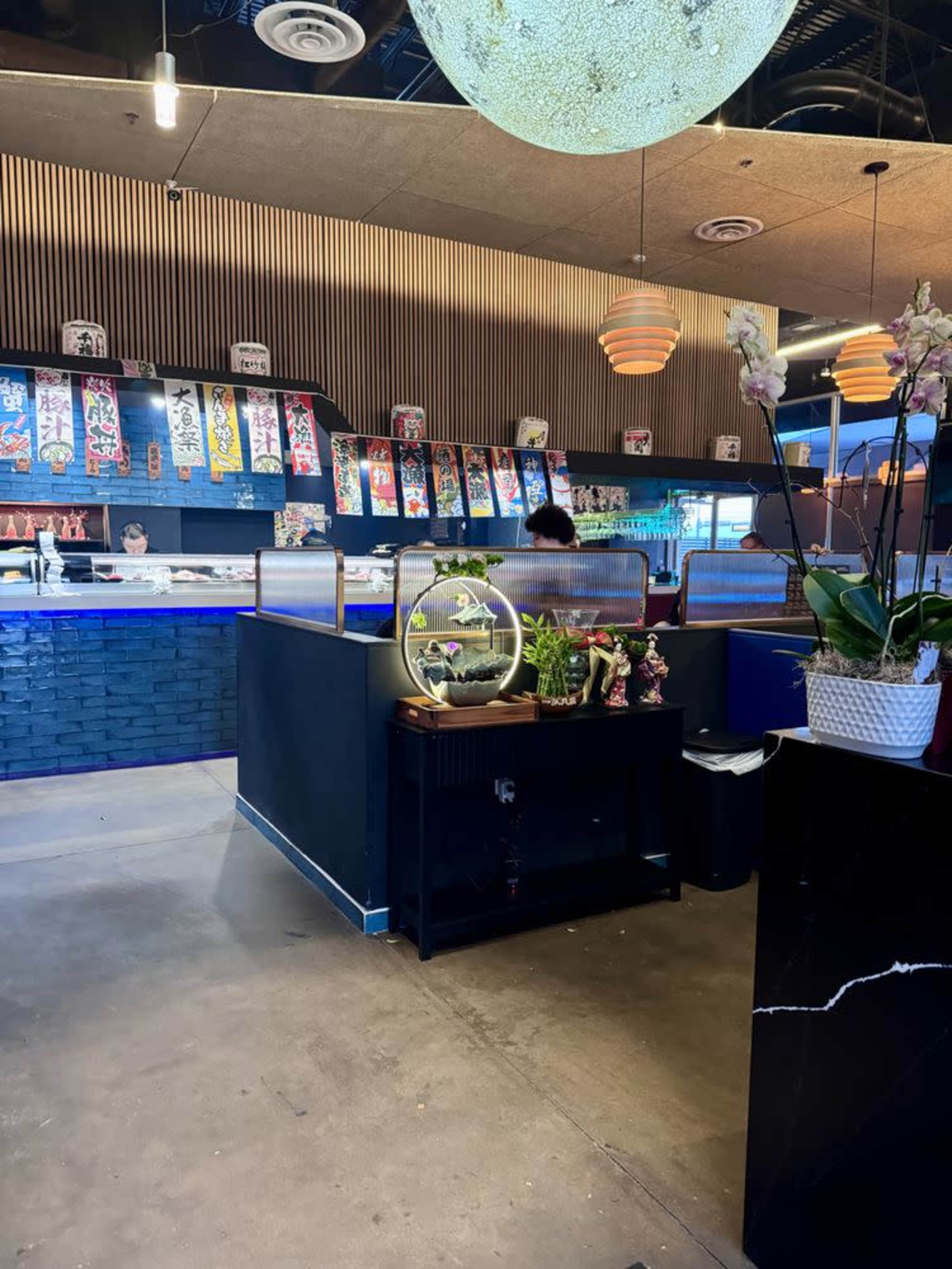 The image shows a modern restaurant interior featuring a sushi counter, decorative wall art, and ornamental plants.