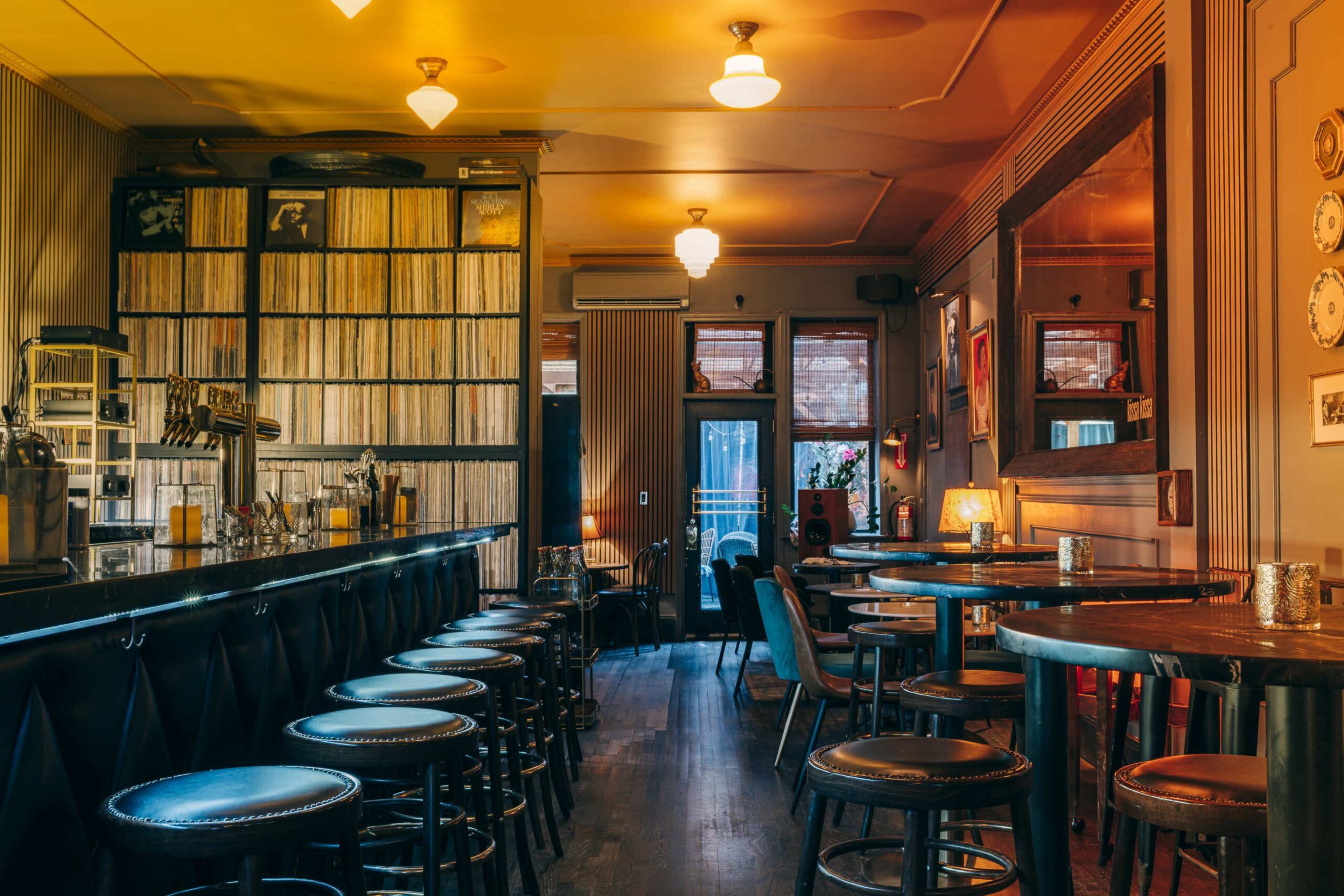 The image shows a dimly lit bar interior with a long bar counter, several round tables, and walls lined with vinyl records.