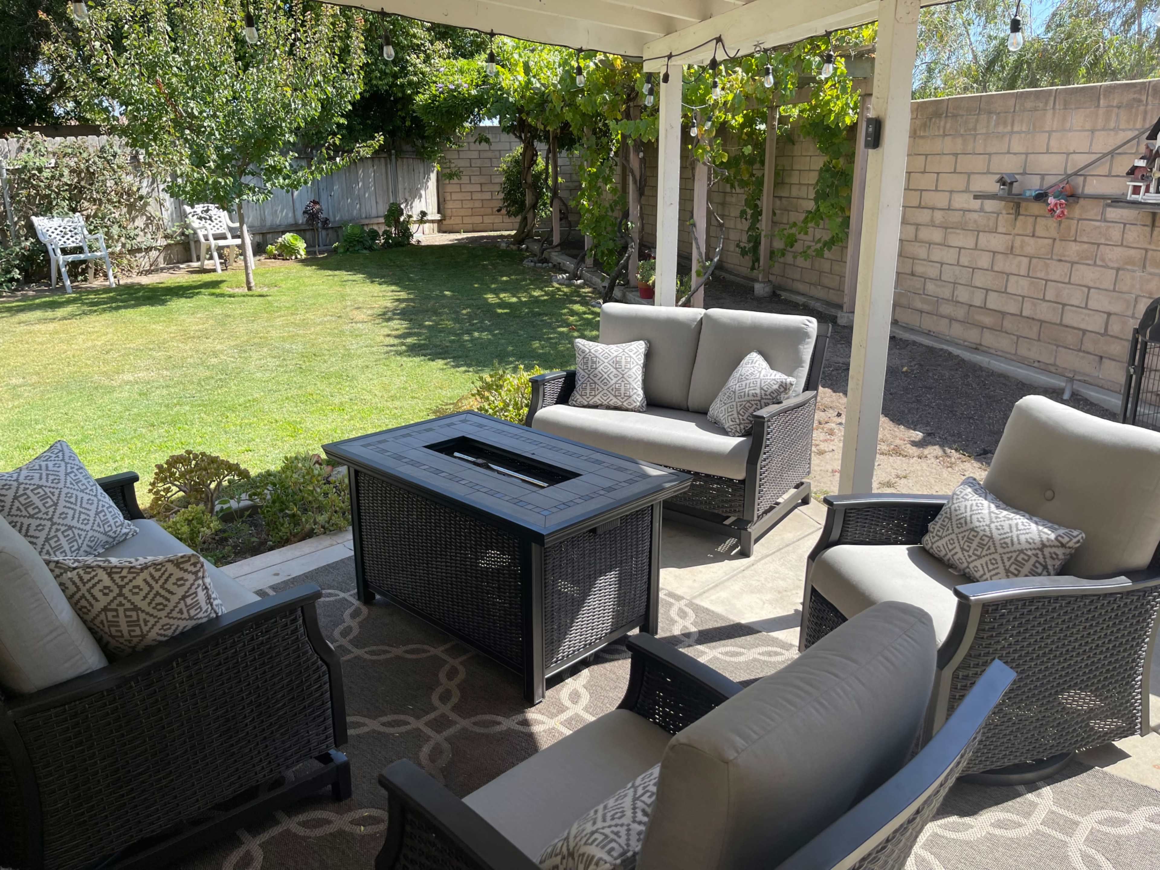 A covered patio with four cushioned chairs surrounding a rectangular table in a backyard featuring grass and a wall of vines.