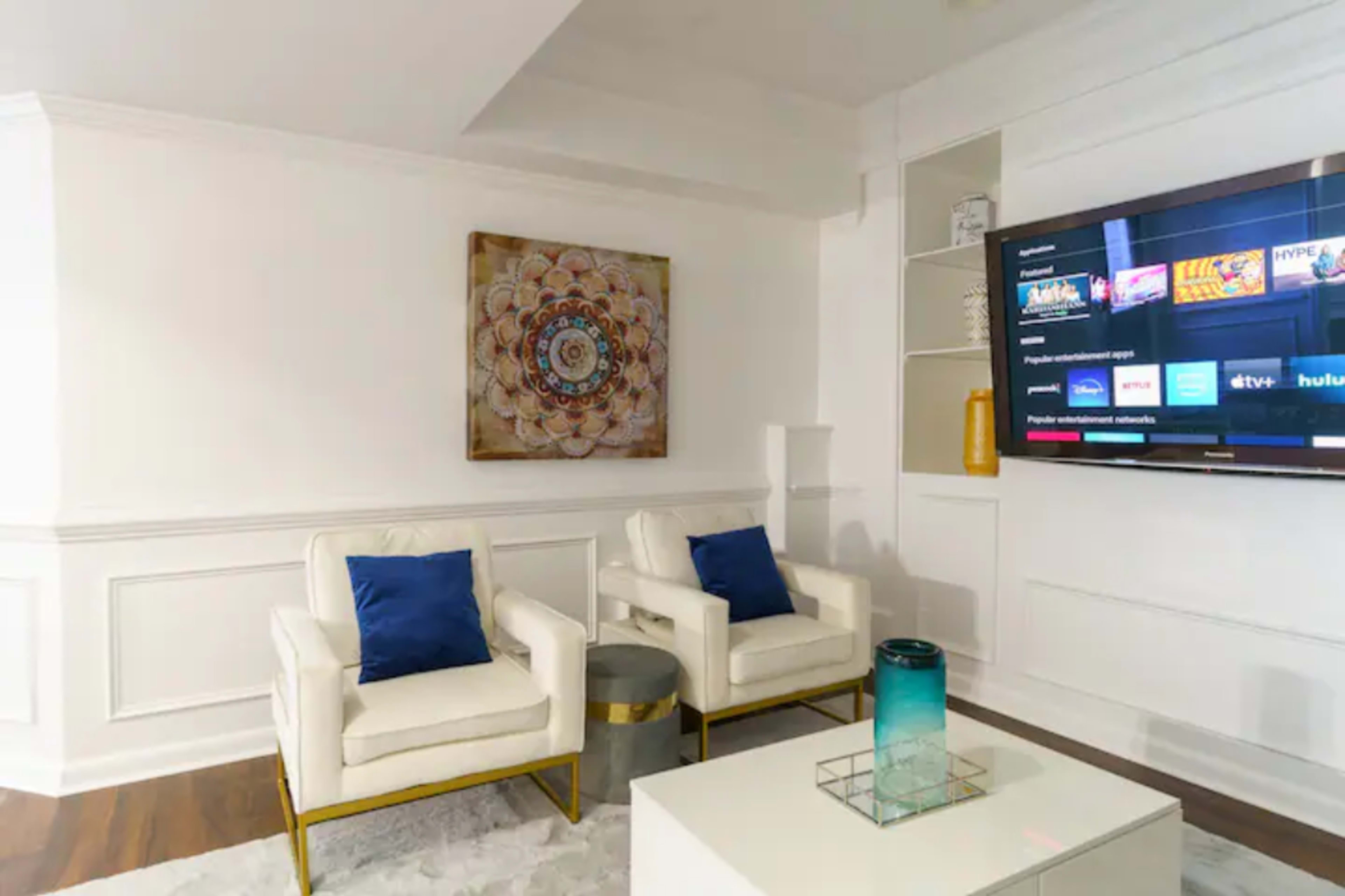 The scene features two white armchairs with blue cushions, a small metallic table, a white coffee table, a turquoise glass vase, and a television mounted on the wall displaying various streaming services, all set against a white wall with decorative paneling.
