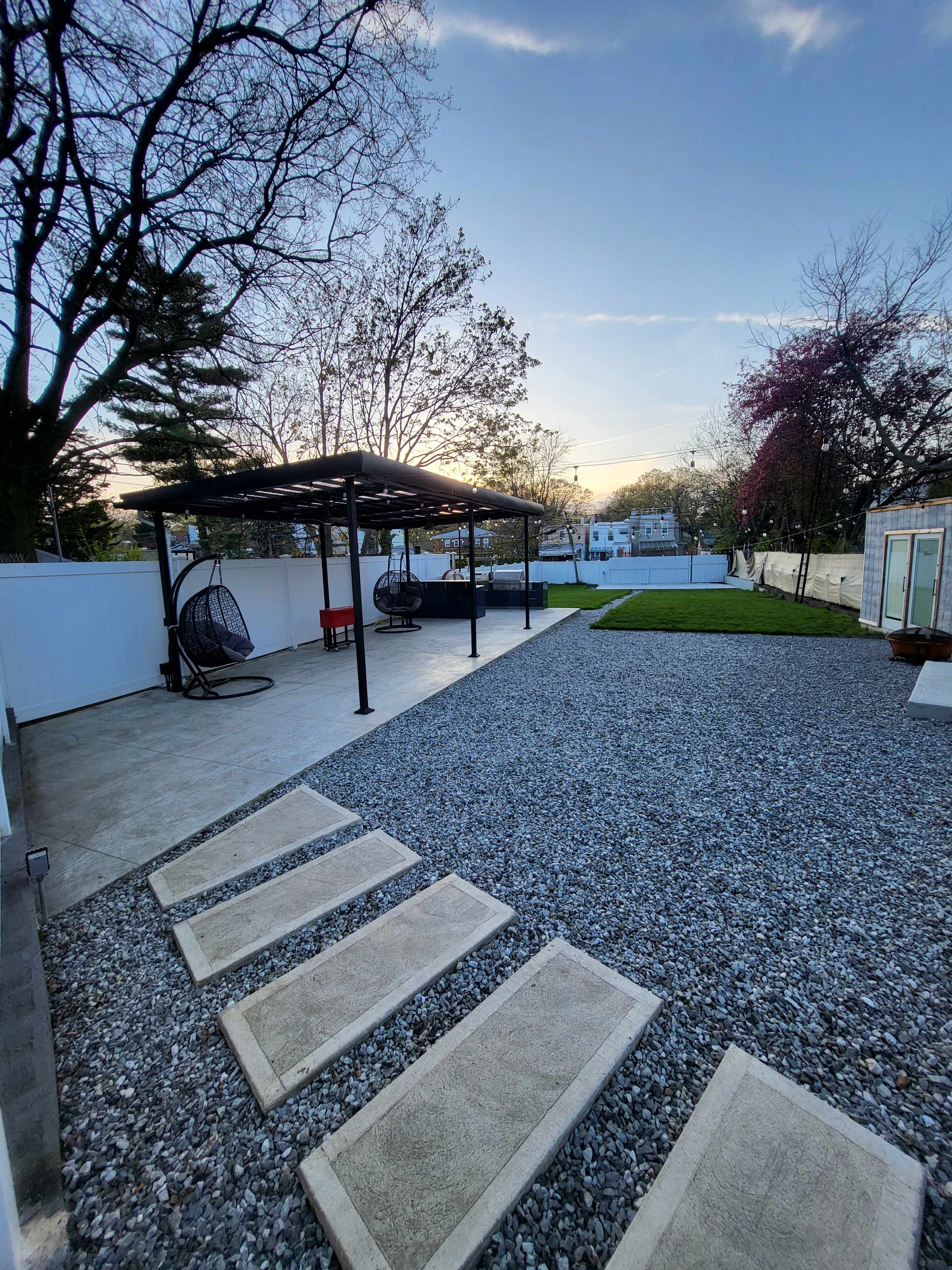 A modern outdoor space with a gravel area, stone pathway, and two hanging chairs under a black pergola, surrounded by greenery and a clear sky.
