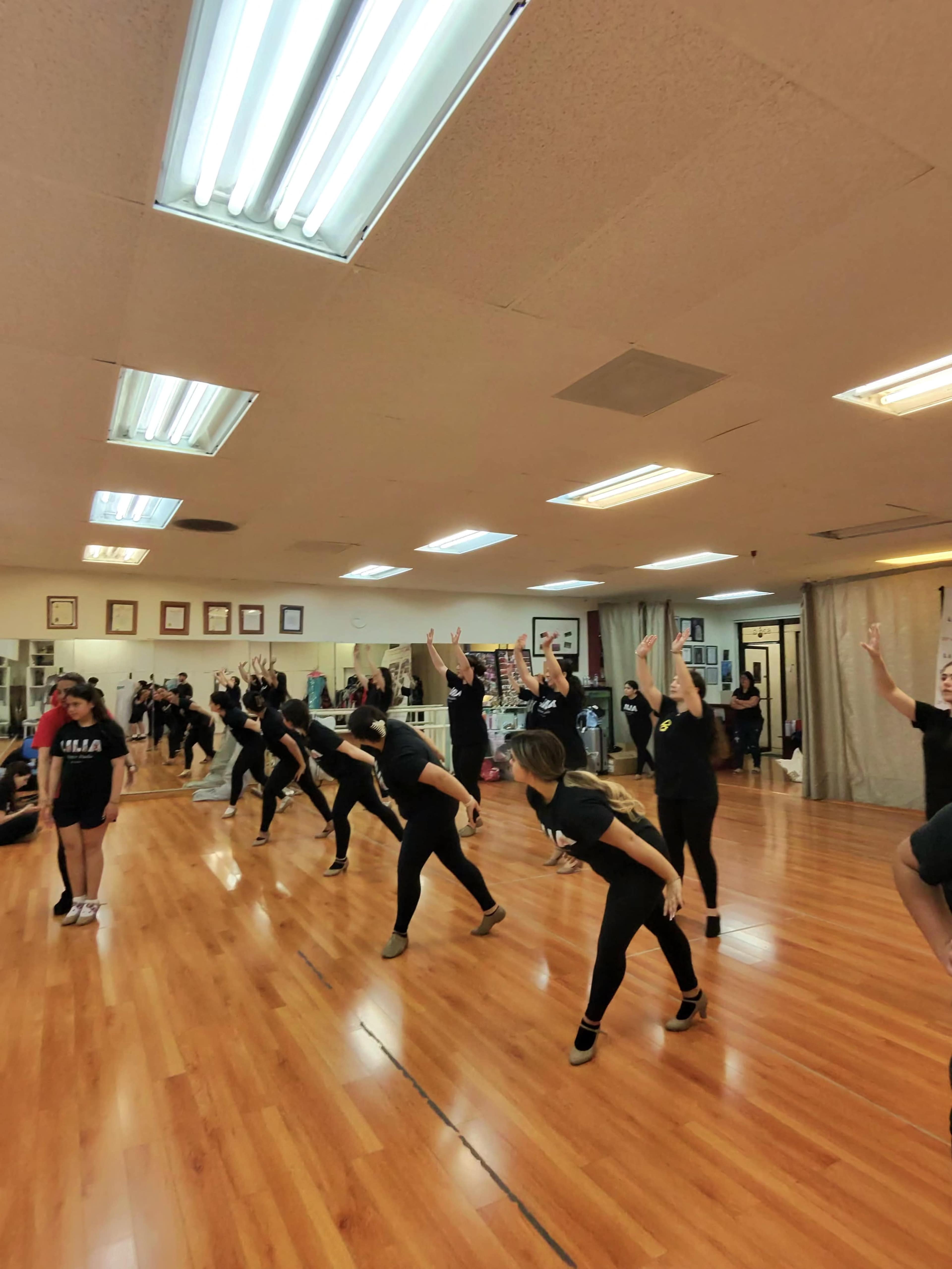 A group of dancers in black attire practice choreography in a spacious dance studio with wooden flooring and bright overhead lights.