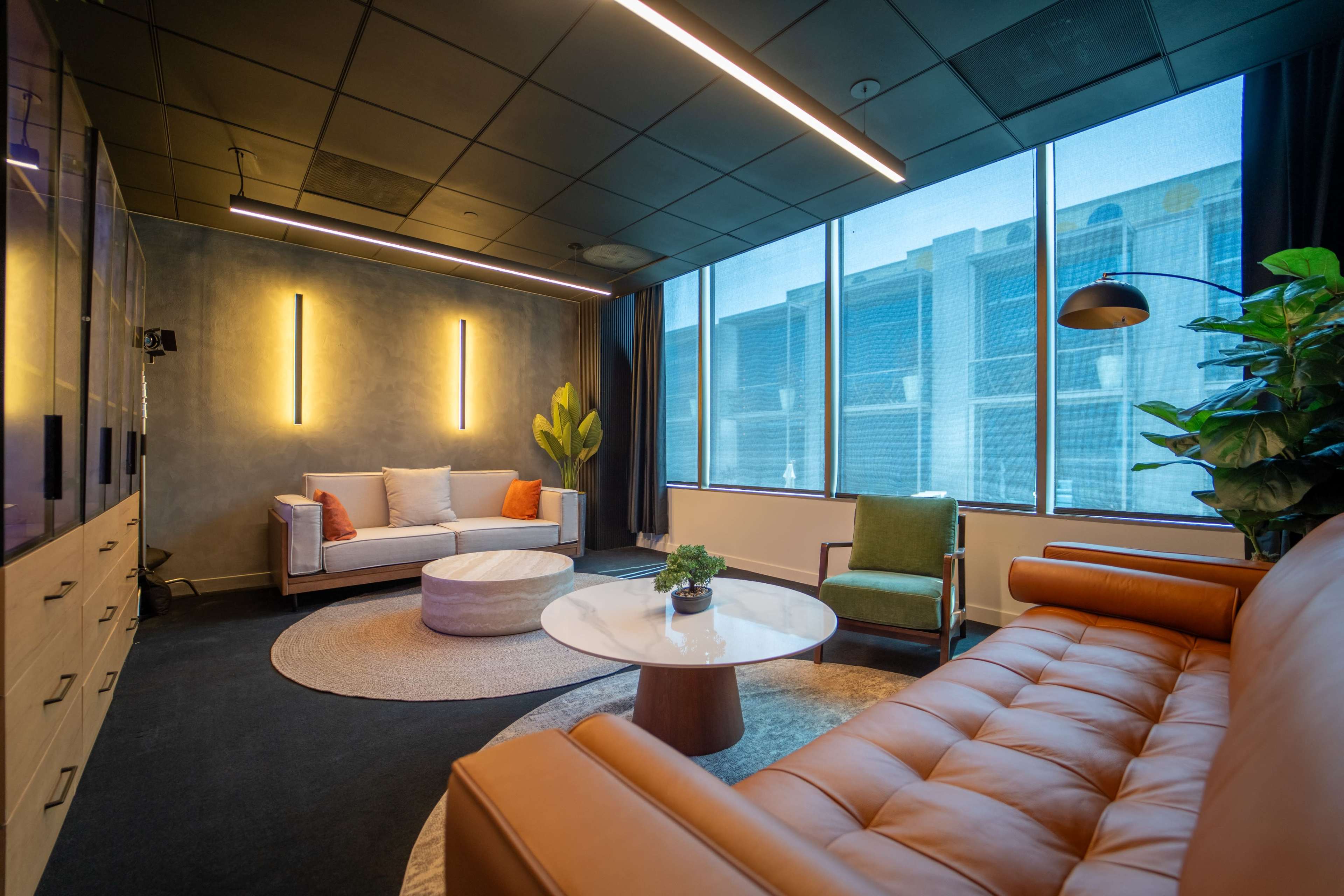 The image shows a modern lounge area featuring a leather sofa, a light-colored couch, a round coffee table, and large windows letting in natural light.