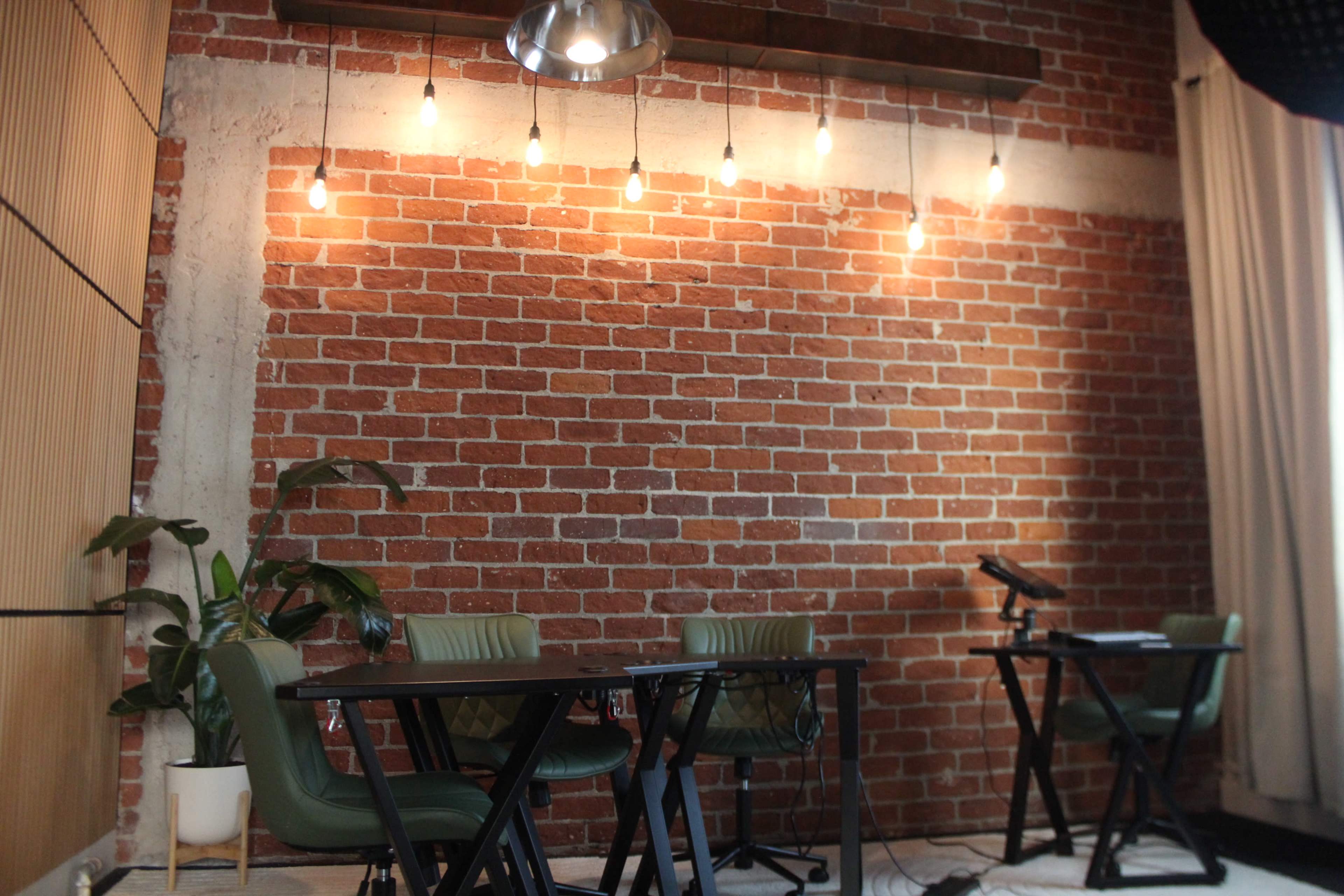 The image shows a room with exposed brick walls, three green chairs around a black table, and warm pendant lights hanging above.