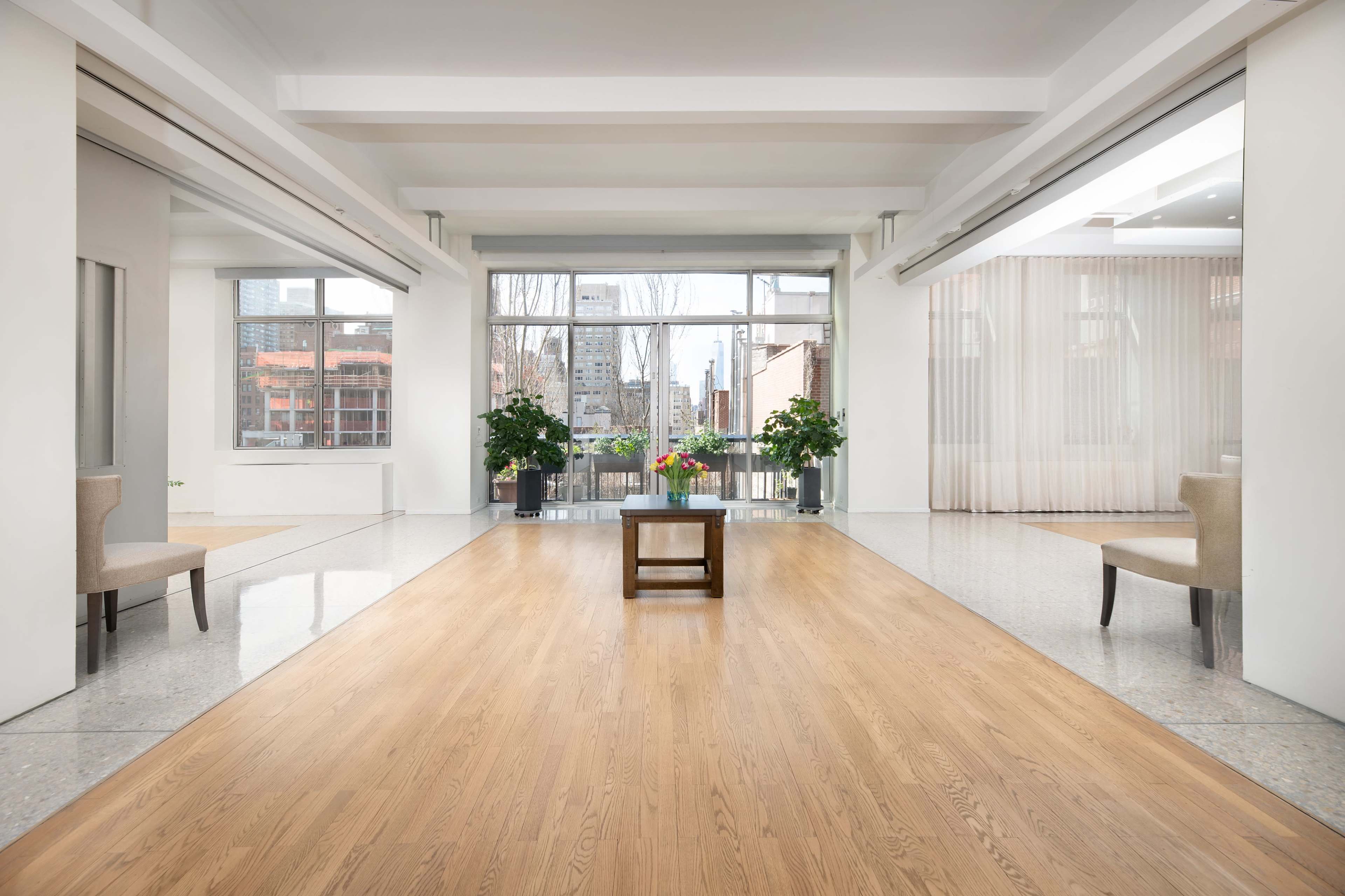 A spacious room with hardwood floors and large windows features two chairs and a small table, with planters and city buildings visible outside.