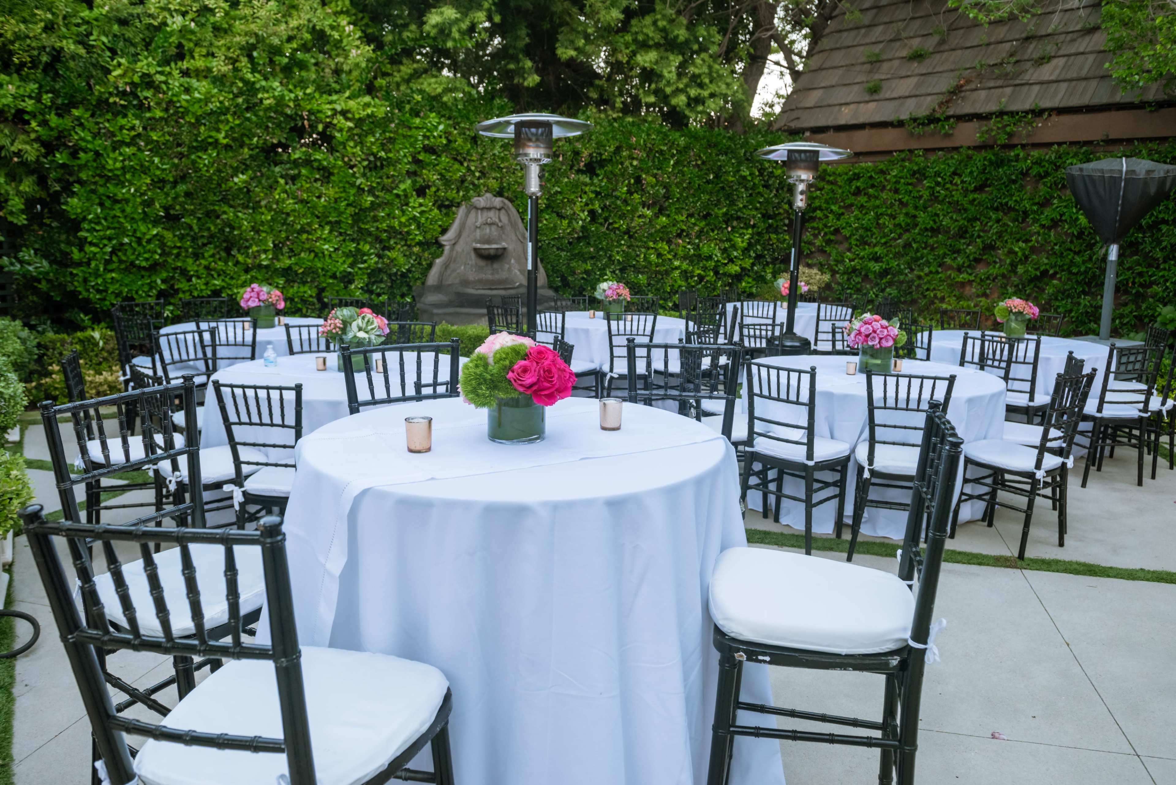 A garden setting features round tables covered with white tablecloths, surrounded by black chairs, and adorned with centerpieces of flowers.