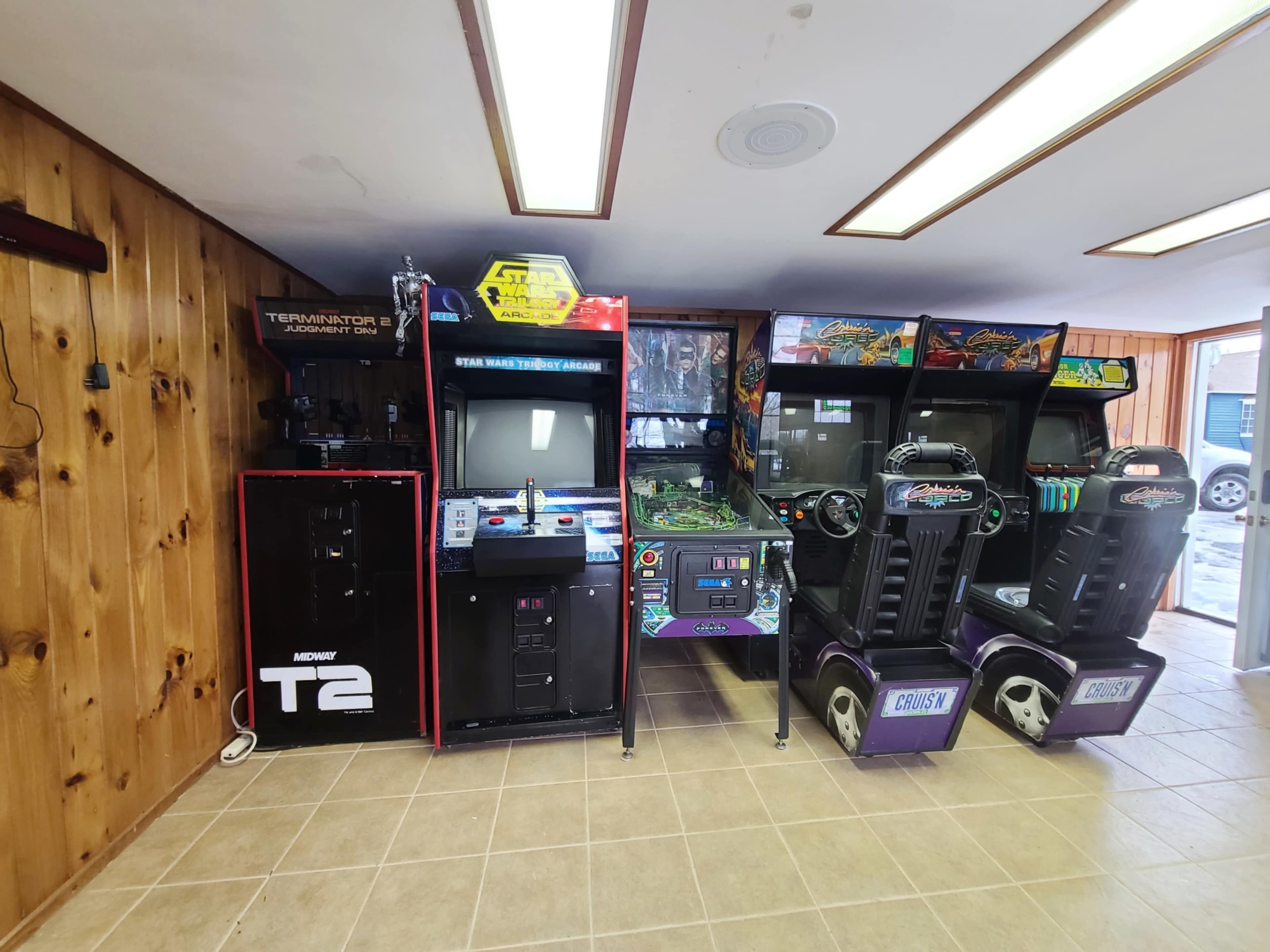 A row of arcade machines, including "Terminator 2," "Star Wars," and racing games, set against a wooden wall in a well-lit room.