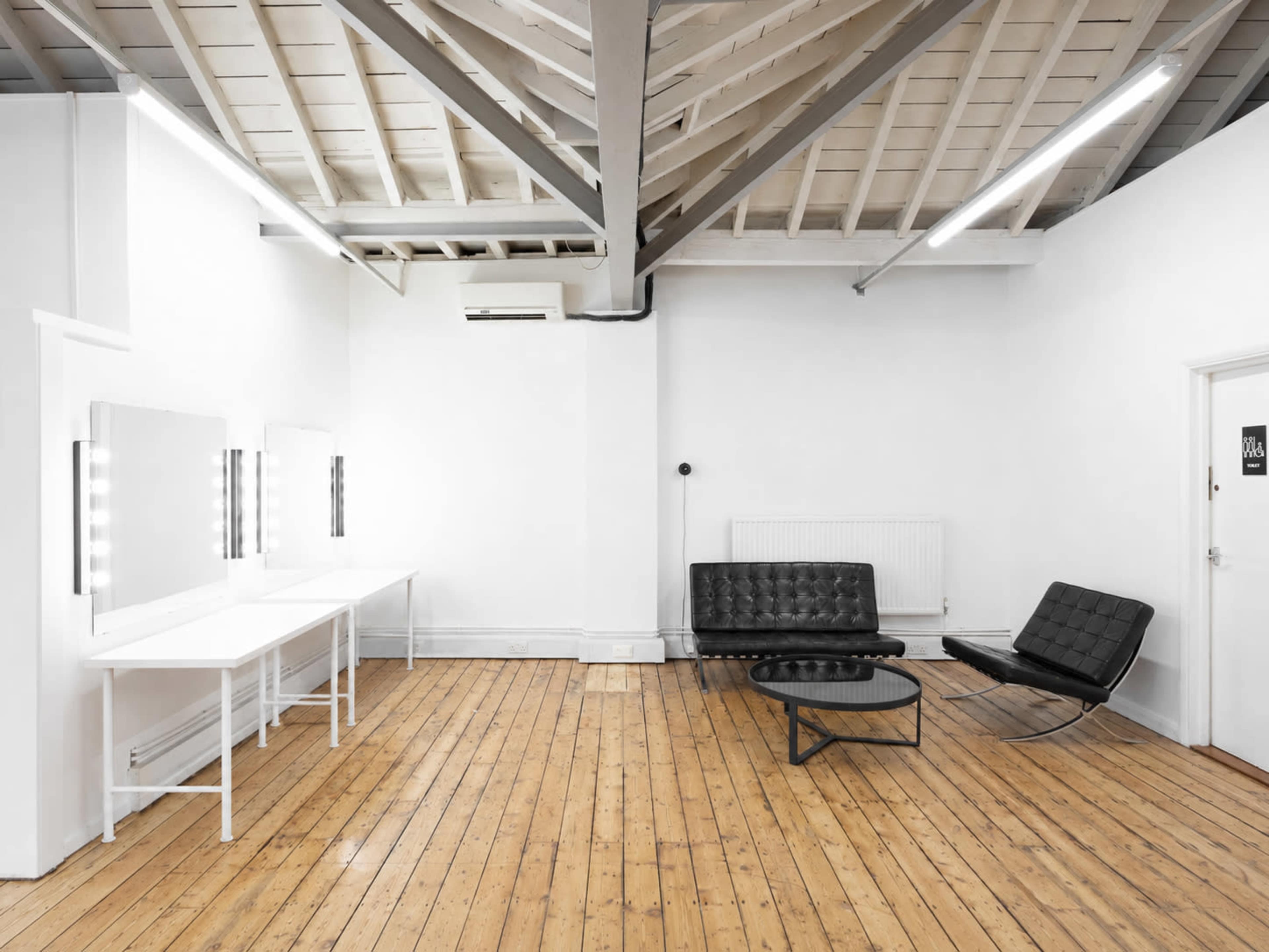 The image shows a minimalistic studio or dressing room with white walls, wooden flooring, a black leather sofa, a round coffee table, and lighting mirrors along one side.