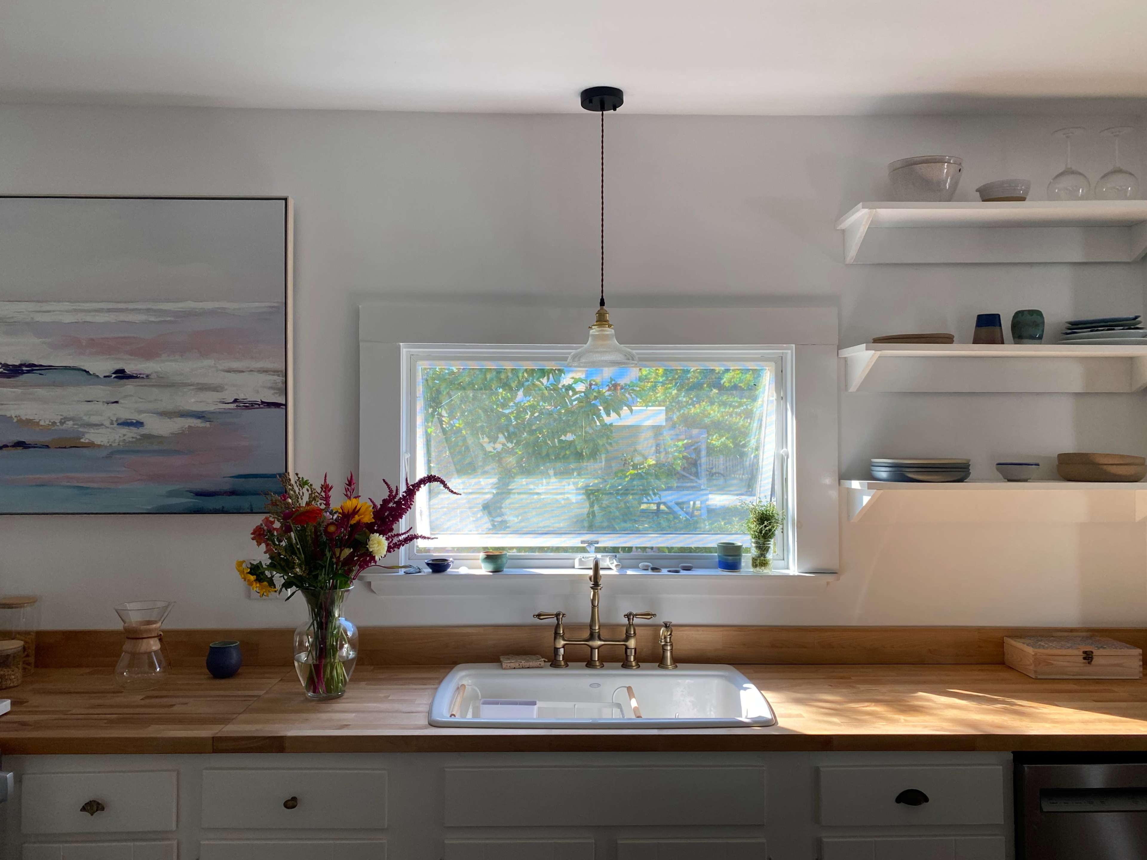 A kitchen features a window above a sink, surrounded by shelves with dishes and a bouquet of flowers on the countertop.