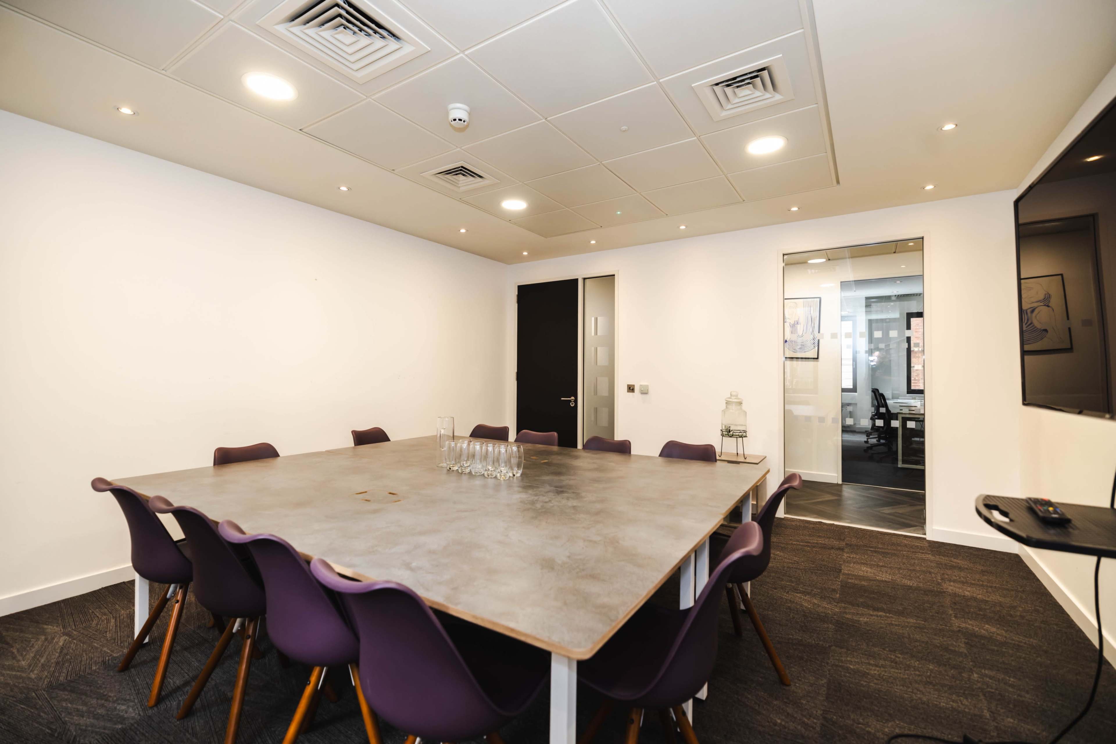 A modern conference room features a large table surrounded by purple chairs, with glass panels and a television on one wall.