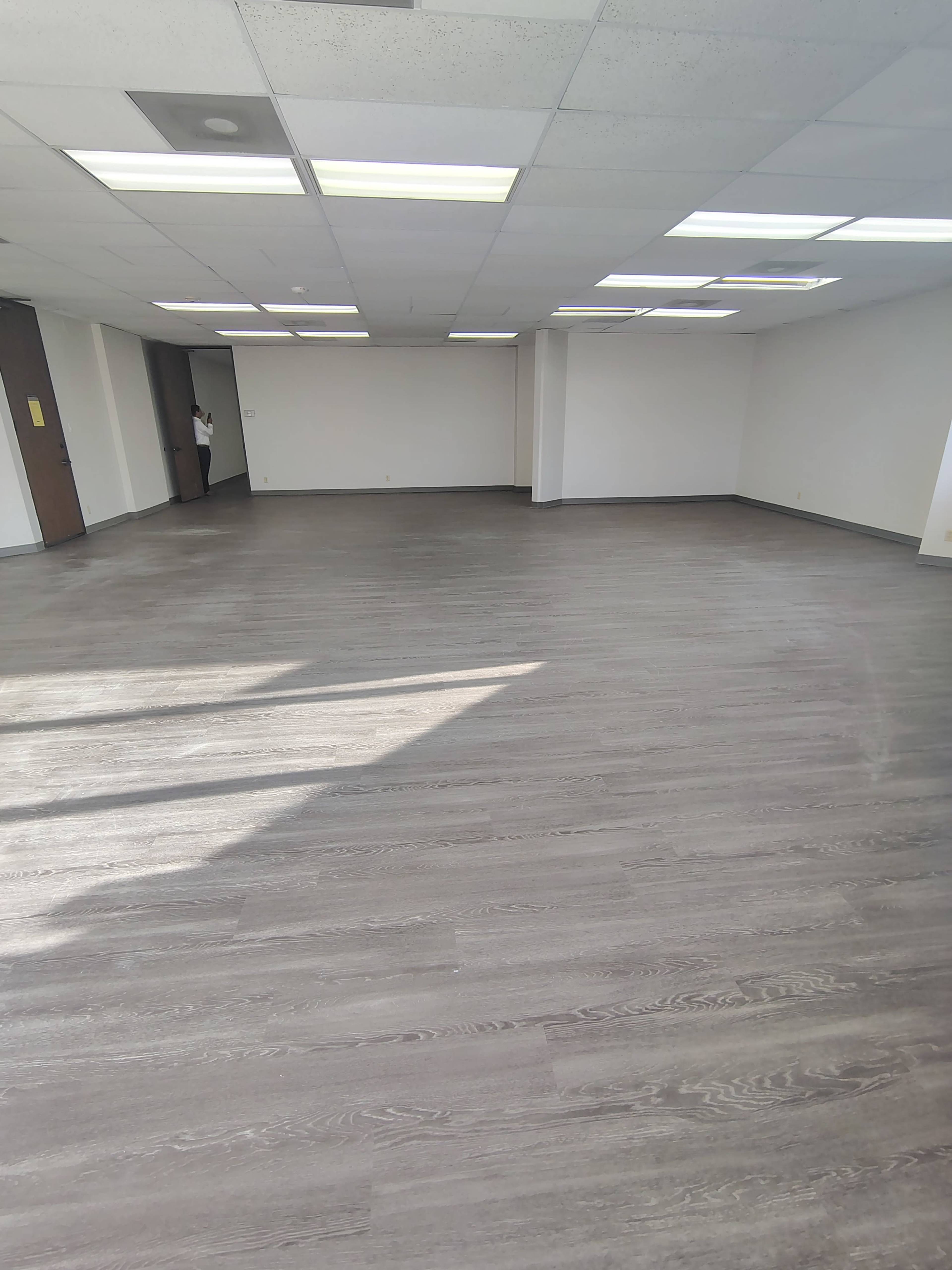 The image shows an empty room with grey wooden flooring and bright overhead lights, featuring white walls and a doorway in the background.