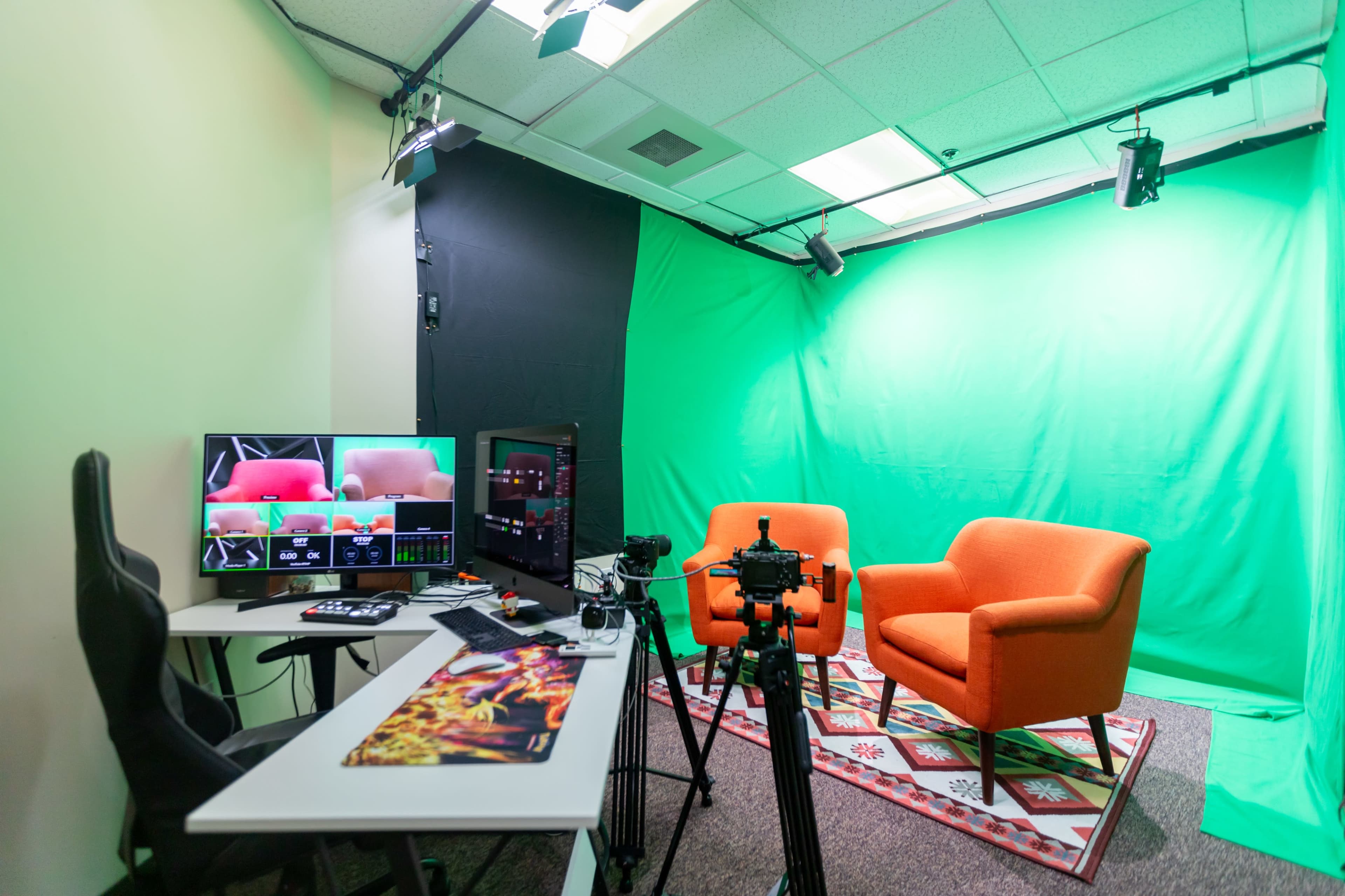 A recording studio featuring two orange chairs on a patterned rug, a computer setup with multiple screens, and a green screen backdrop.