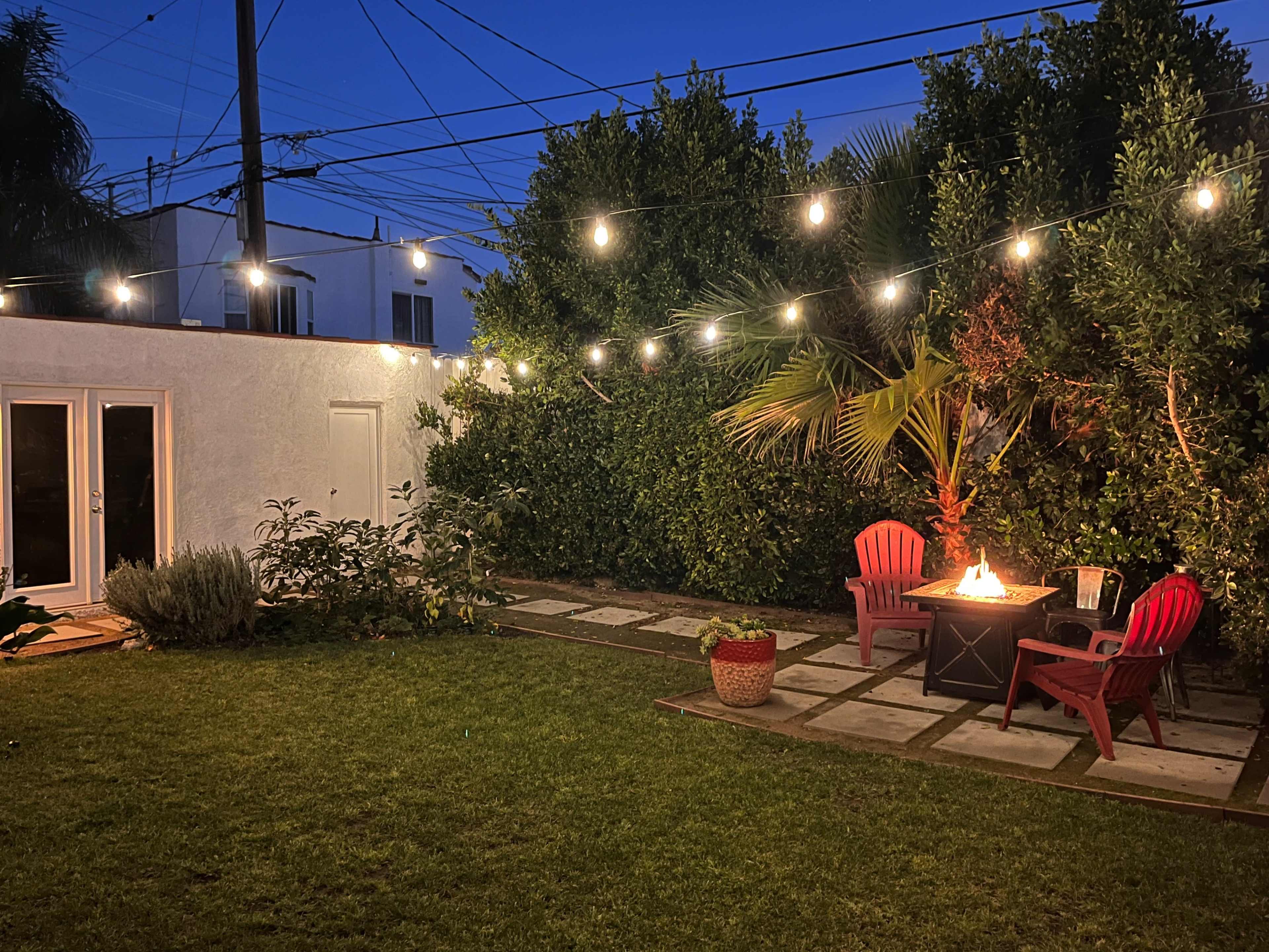 A backyard scene features a fire pit surrounded by red chairs, illuminated by string lights, with greenery and a house in the background.
