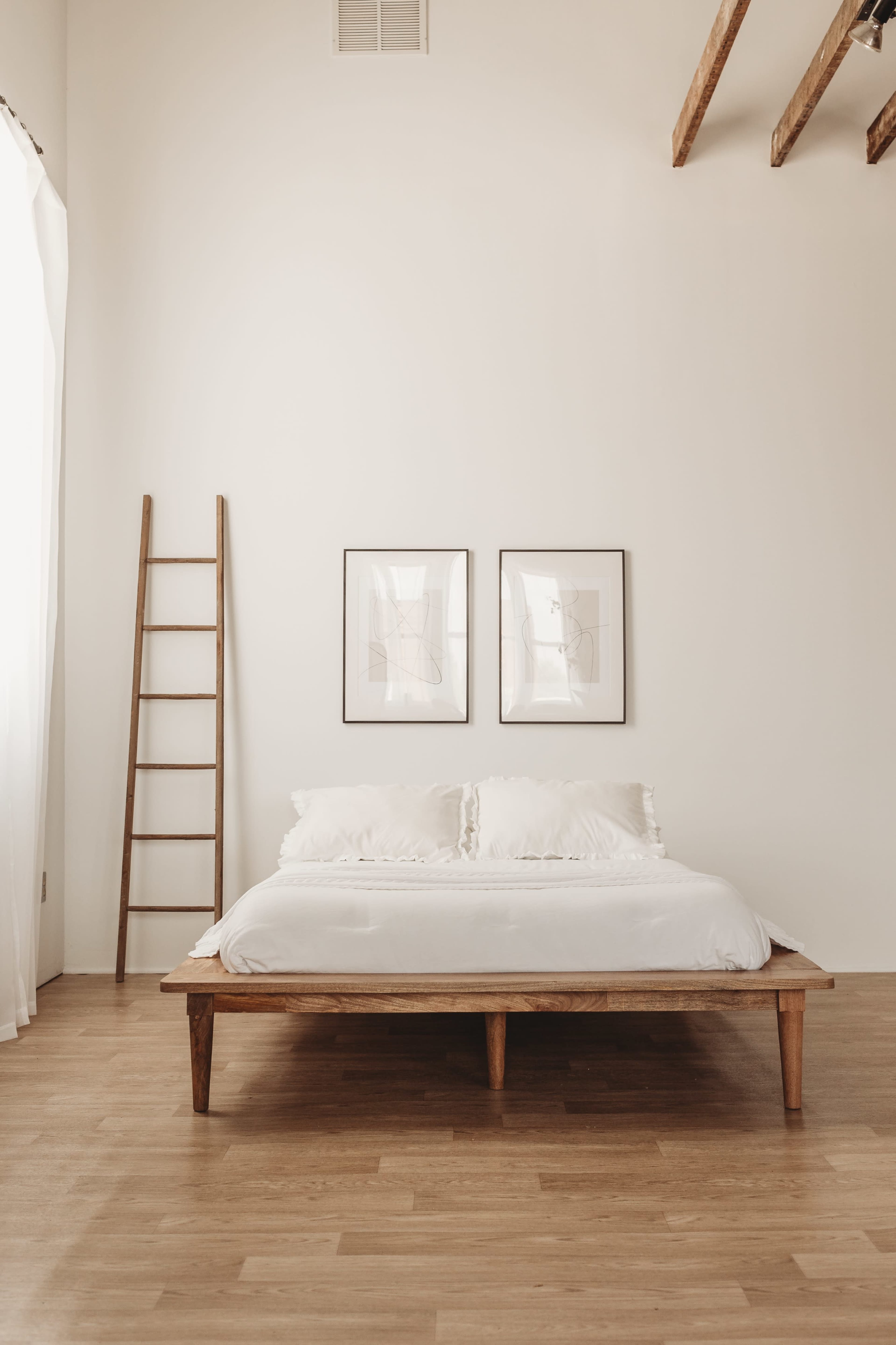 A minimalist bedroom features a wooden bed frame with a white bedding set, two framed artworks on the wall, and a wooden ladder leaning against the wall.