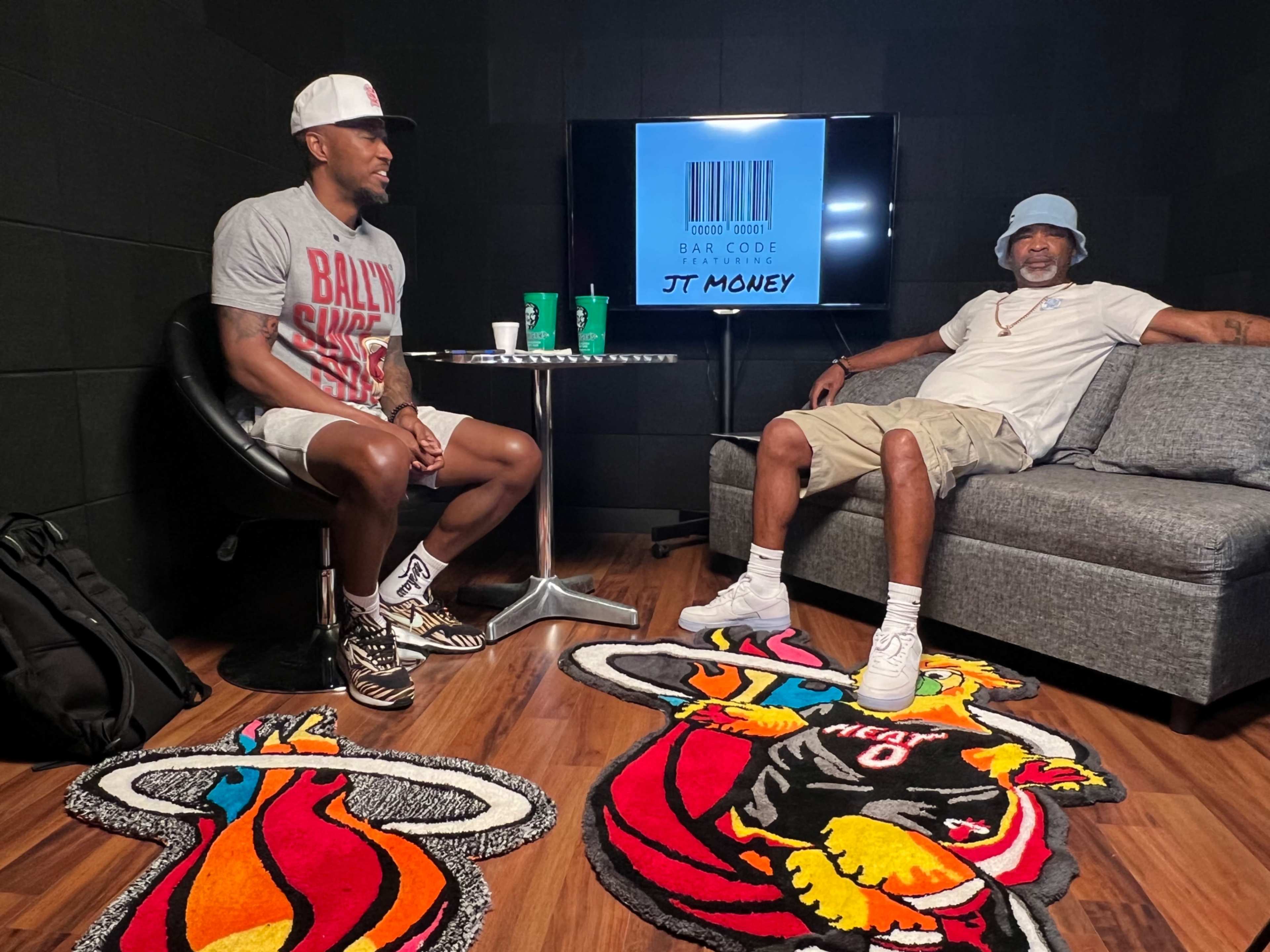 Two men sit in a recording studio setting, one at a table and the other on a couch, with Miami Heat-themed rugs on the floor and a display featuring "JT MONEY" in the background.