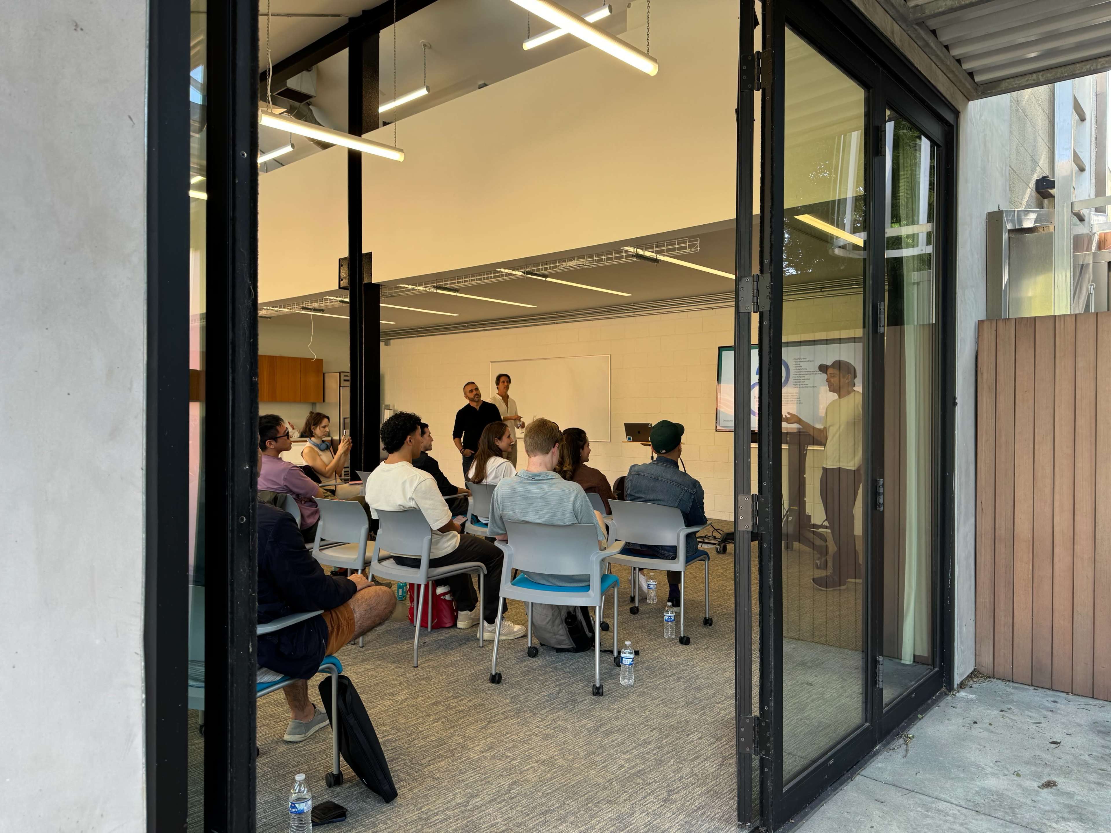 A group of people is seated in a classroom setting, facing two presenters in front of a screen.
