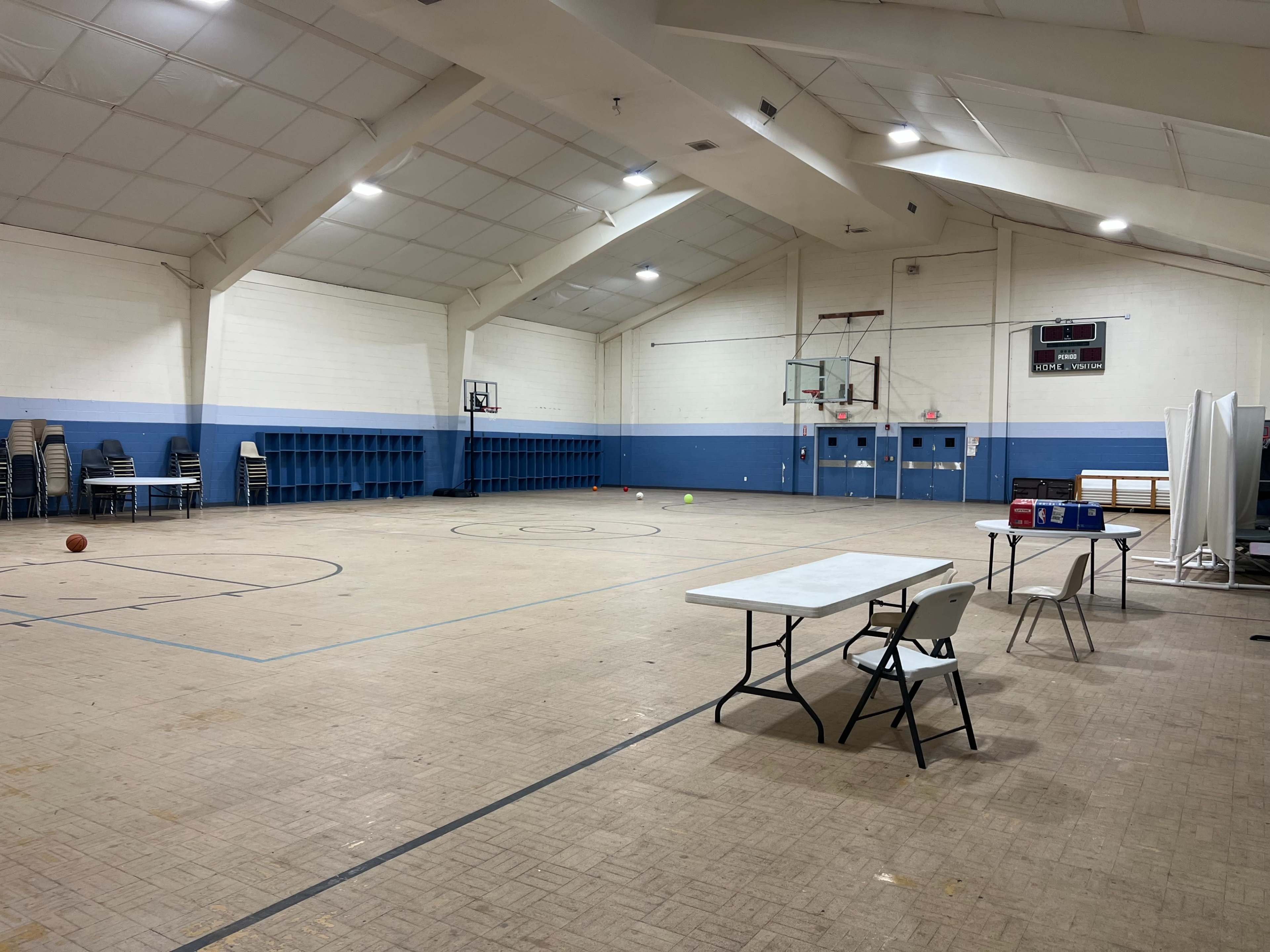 The image shows a spacious indoor gymnasium with basketball hoops, a few scattered balls, and several empty folding chairs and tables along the walls.
