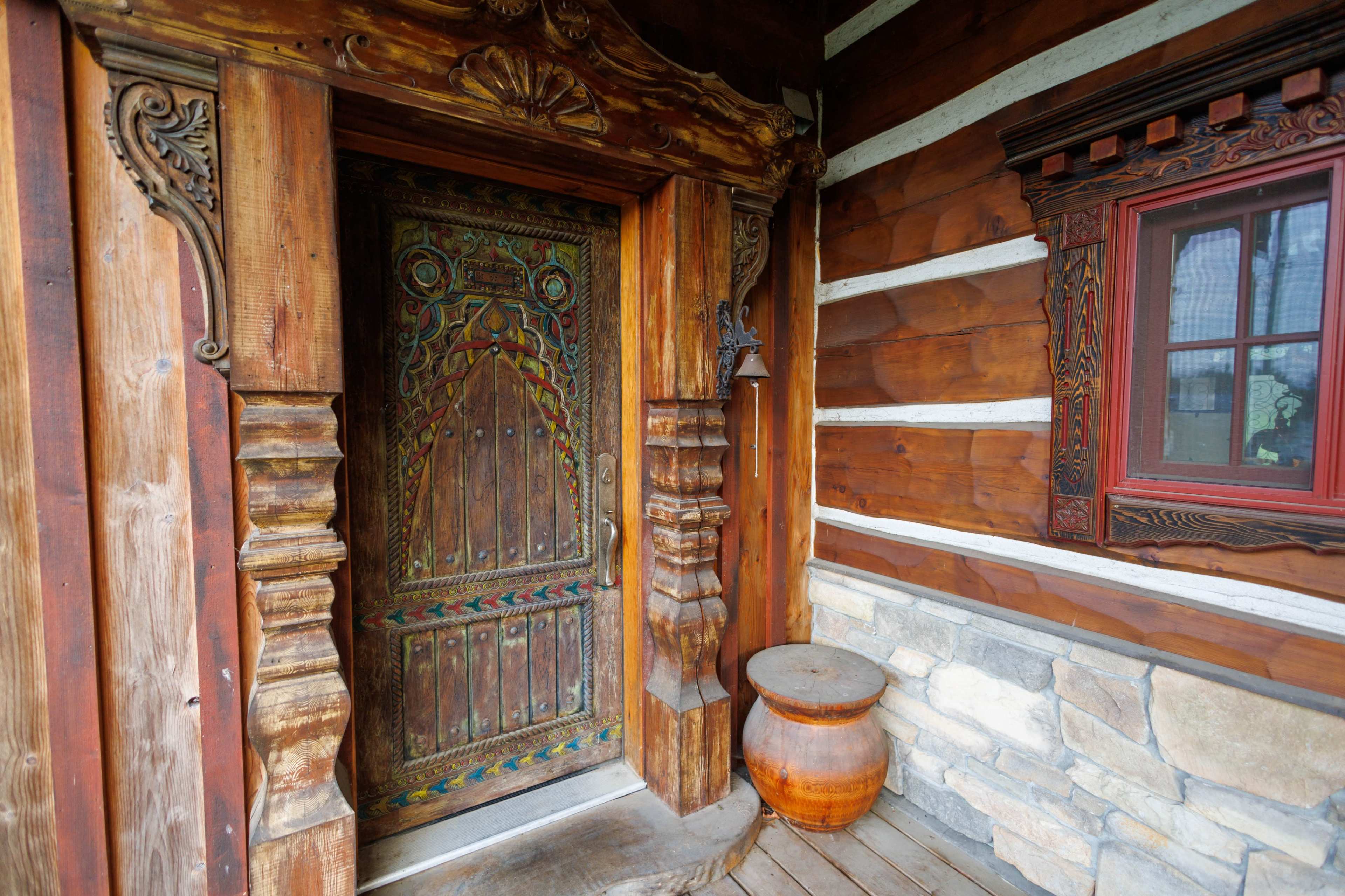 The image shows a rustic wooden door with intricate carvings, framed by log cabin walls and accompanied by a decorative window and a stone base.