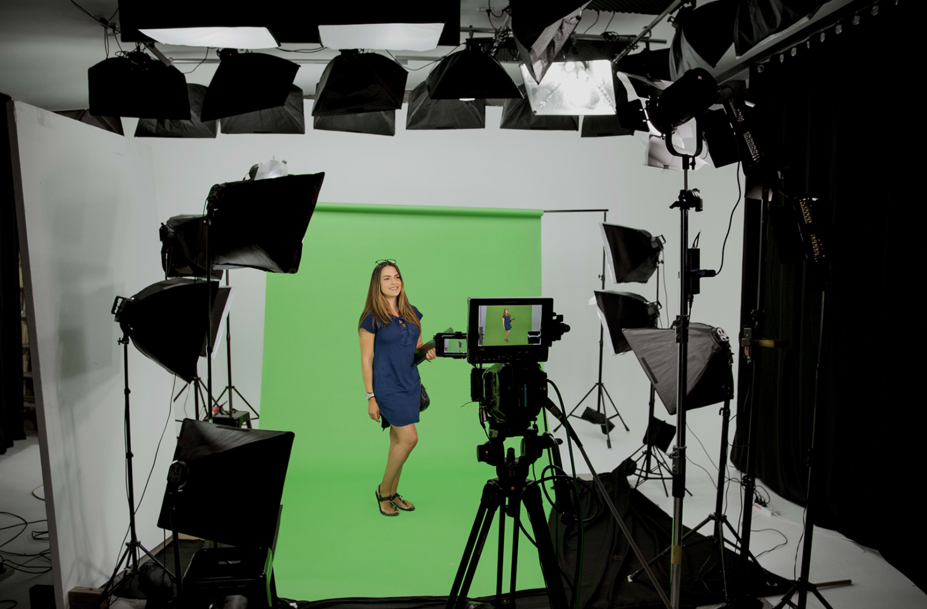 A woman stands on a green screen set surrounded by studio lights and camera equipment.