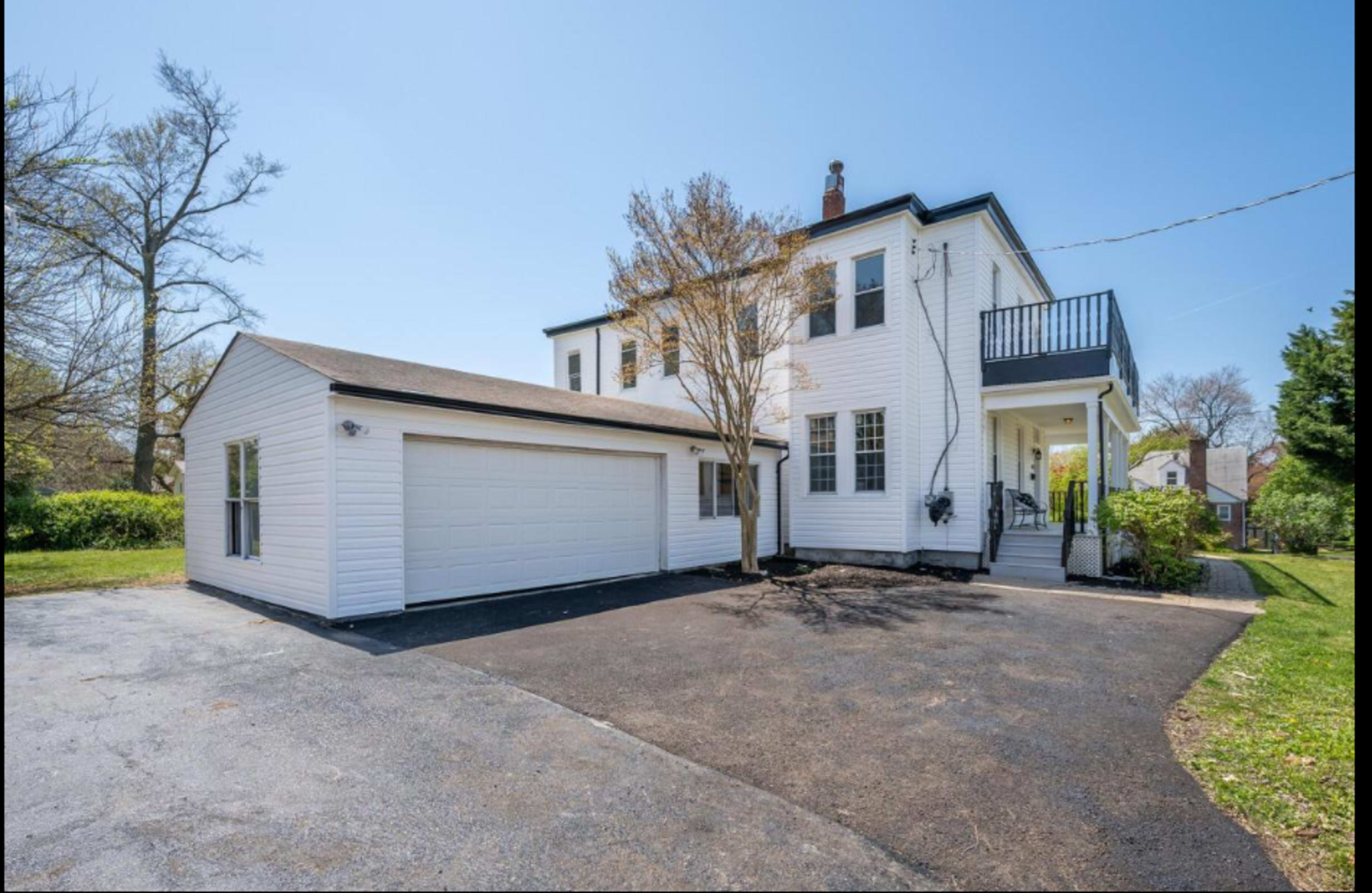 A two-story white house with a garage and balcony is situated on a paved driveway surrounded by greenery.