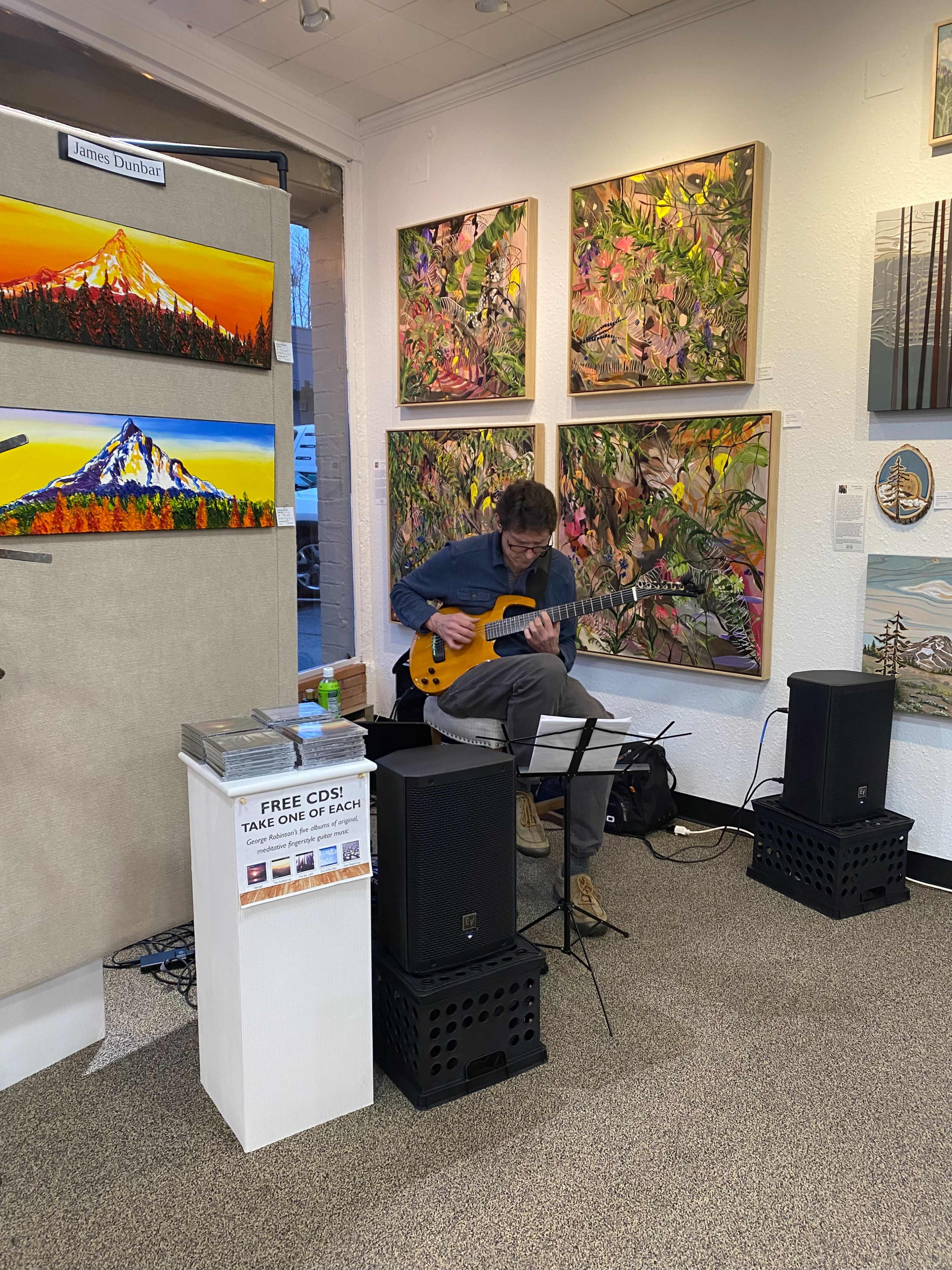 A musician sits on a stool playing an electric guitar in an art gallery filled with colorful paintings.