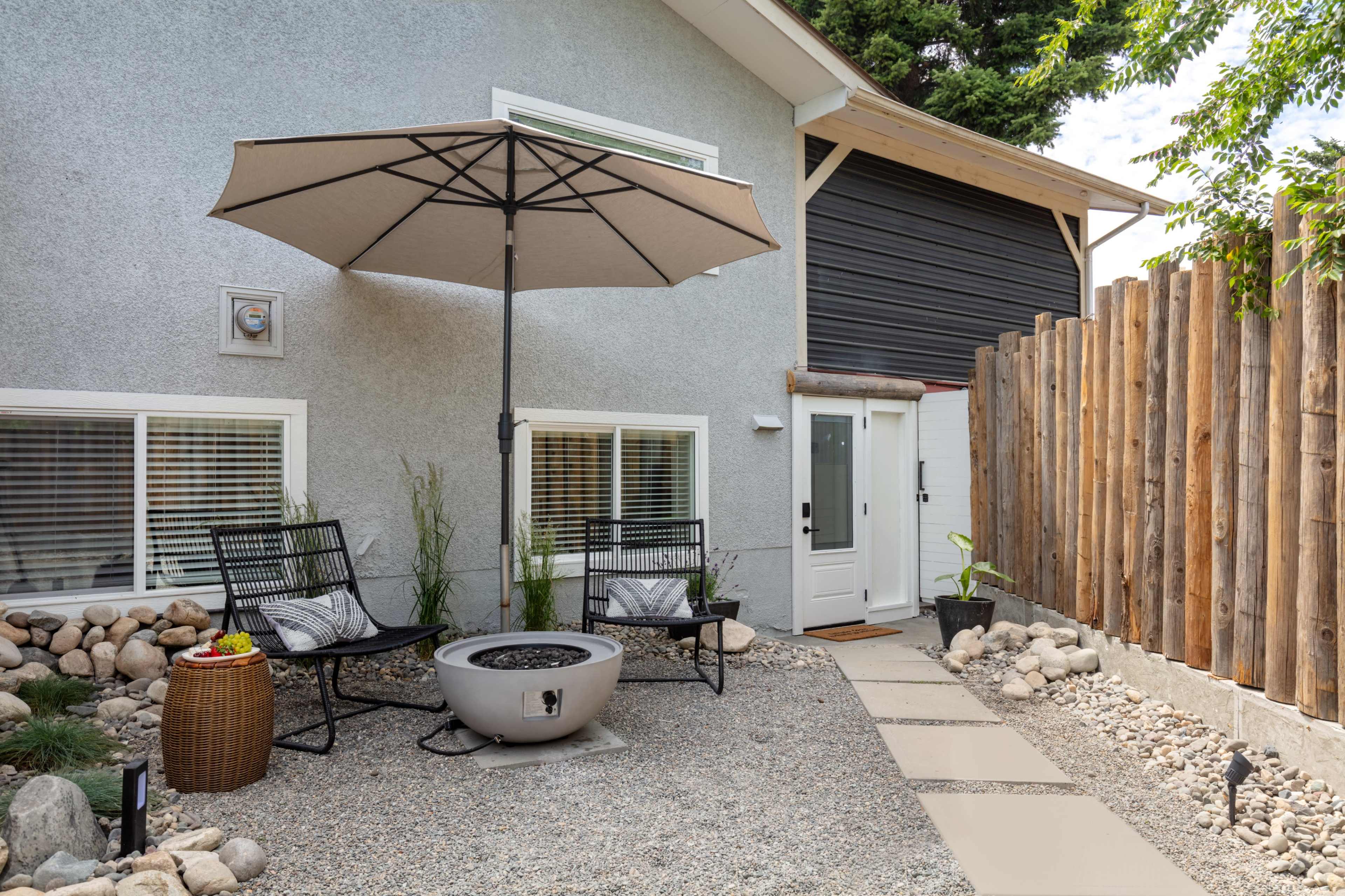 A backyard patio area with two black chairs, a round fire pit, a large umbrella, and a gravel path leading to a house with a wooden fence.