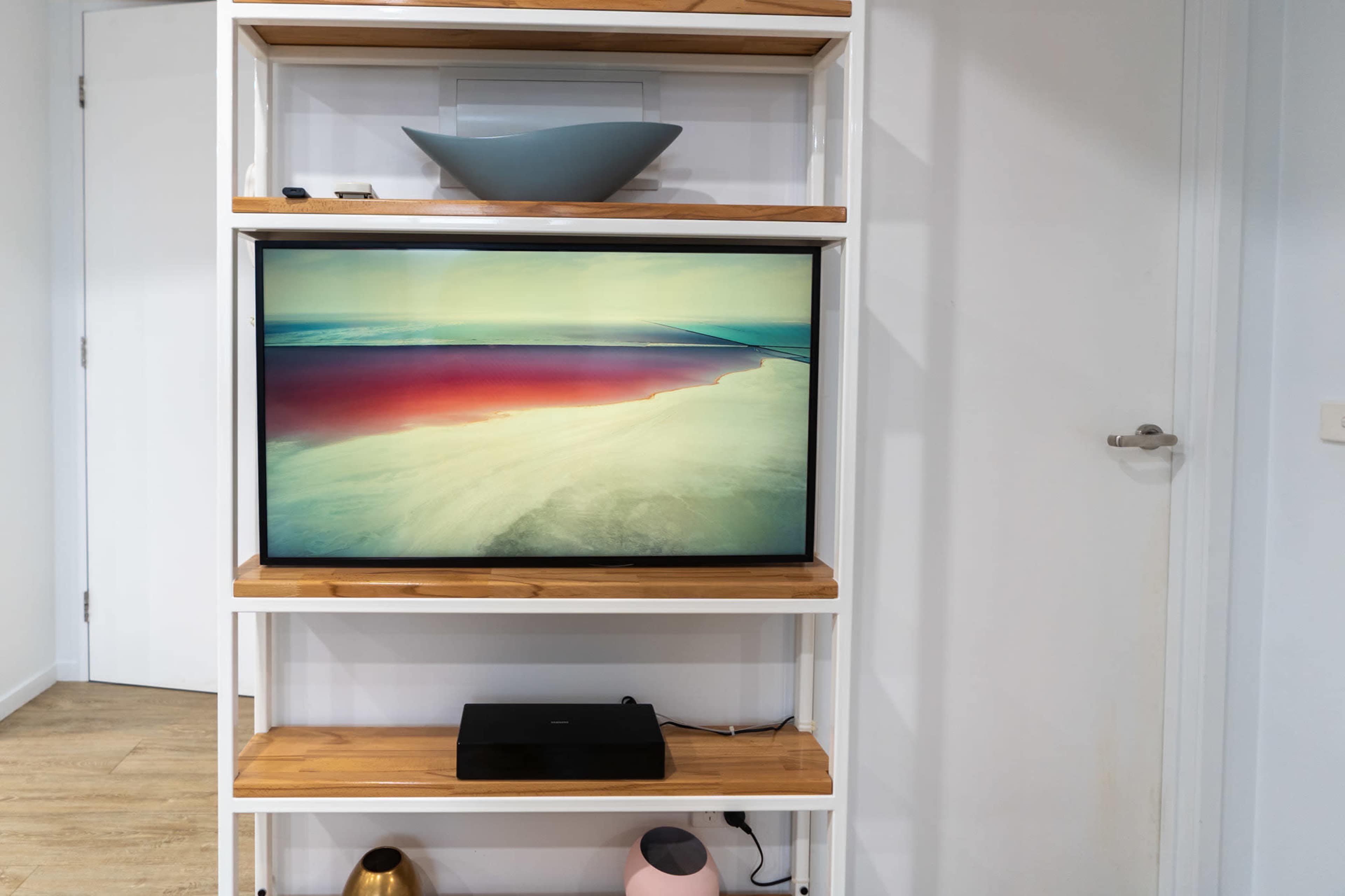 A modern shelving unit displays a television with a landscape image above a media device, accompanied by minimalist decorative items on the upper shelves.