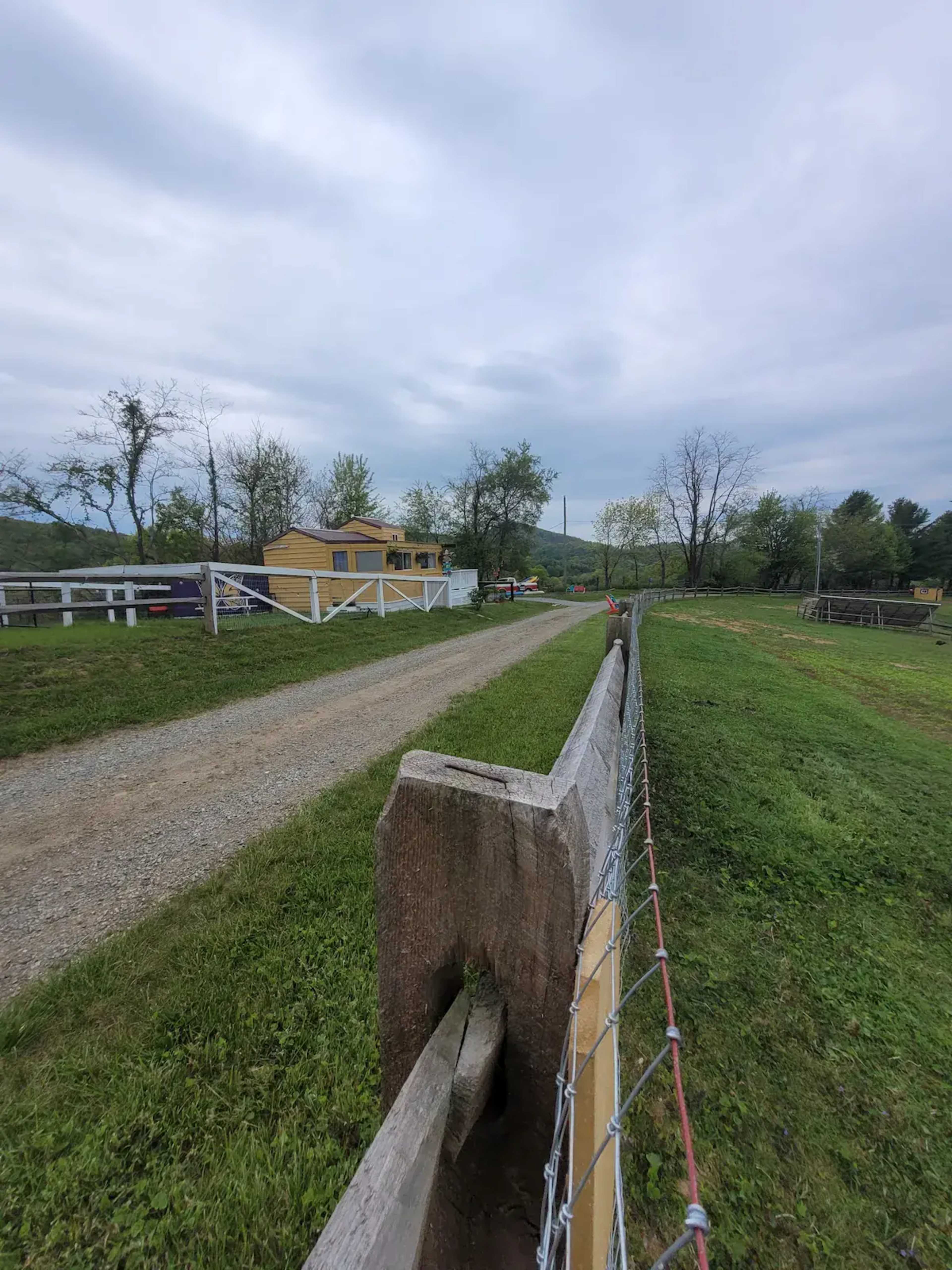 A gravel road leads towards a yellow house surrounded by a grassy landscape and a wire fence.