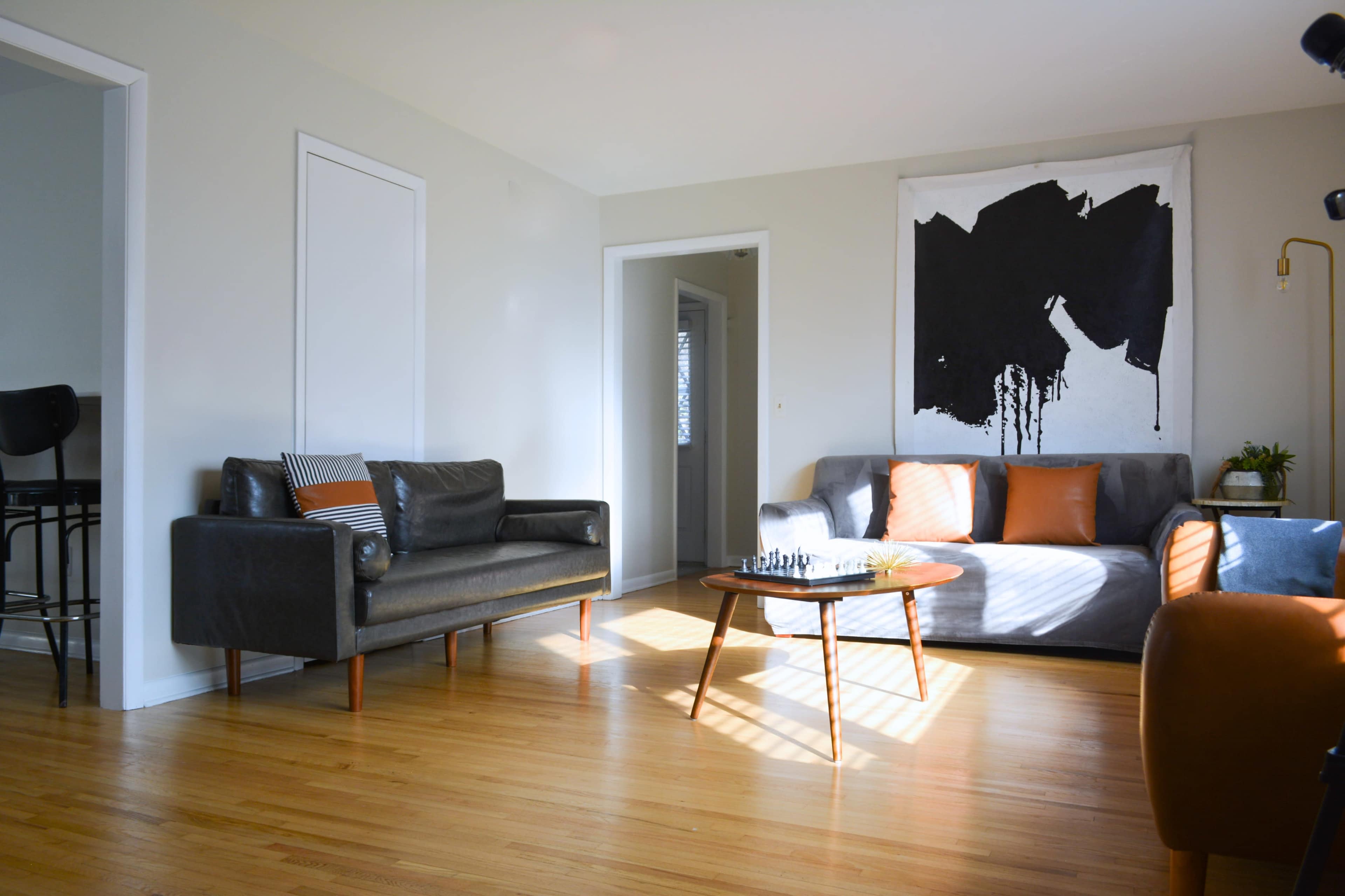 The living room features a gray sofa and a black leather couch, with a wooden coffee table at the center and an abstract wall hanging above.