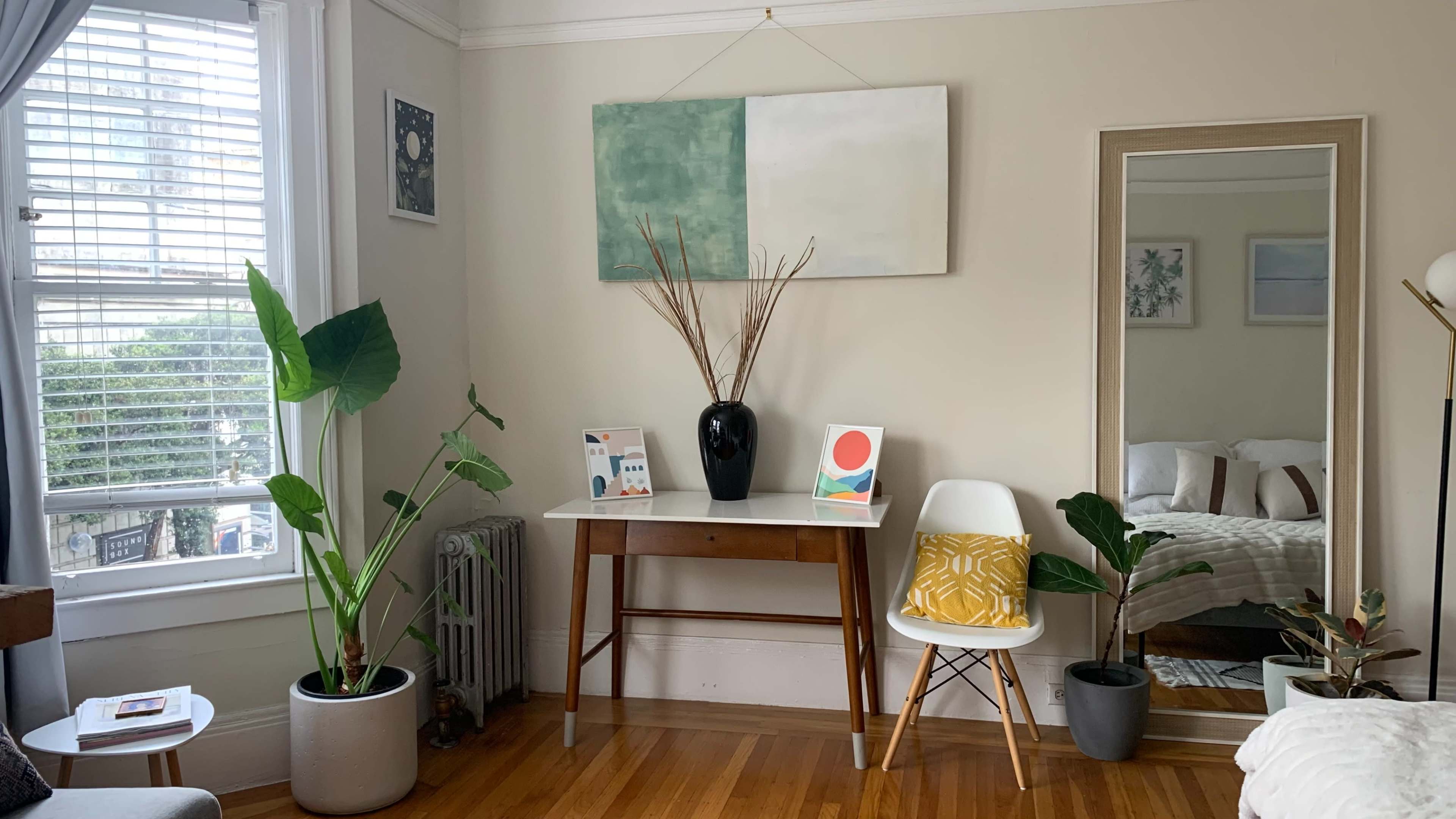 A living room corner featuring a table with a black vase and two art prints, accompanied by potted plants and a mirror against a light-colored wall.