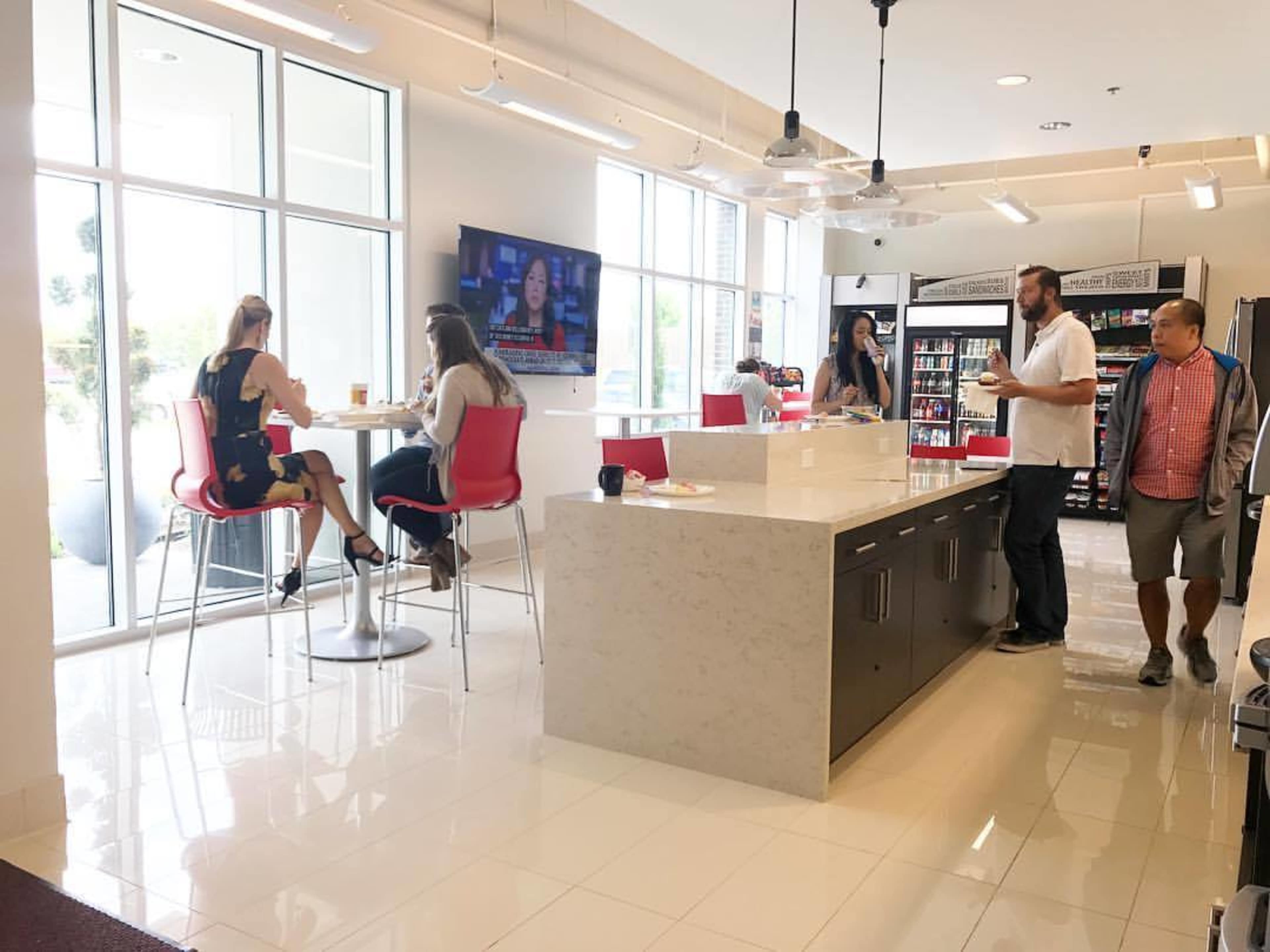 The image shows a modern café area with a white countertop, people sitting at high tables, and a television displaying news in the background.