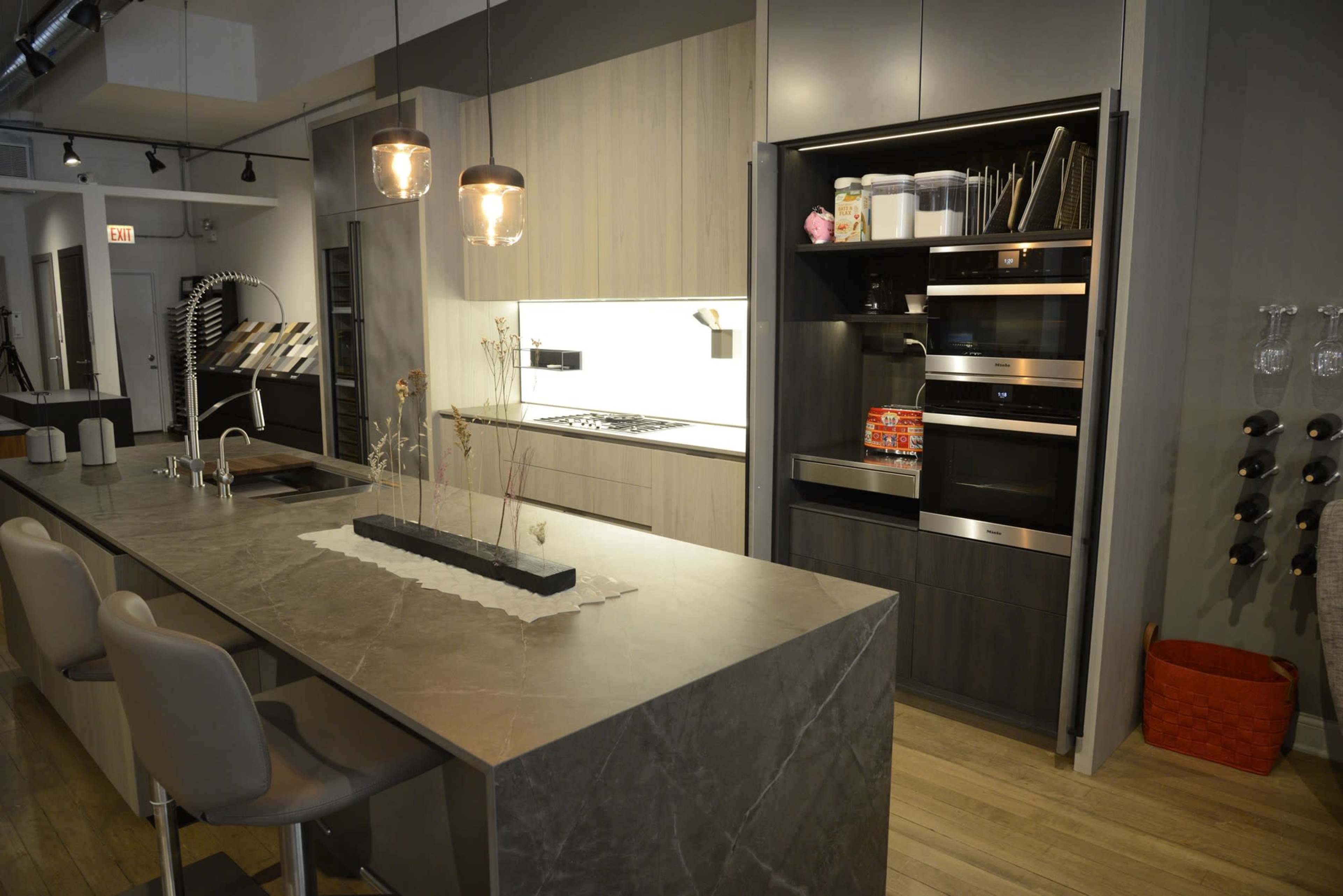 A modern kitchen with a large gray stone countertop, sleek cabinetry, and built-in appliances, along with three light-colored bar stools.