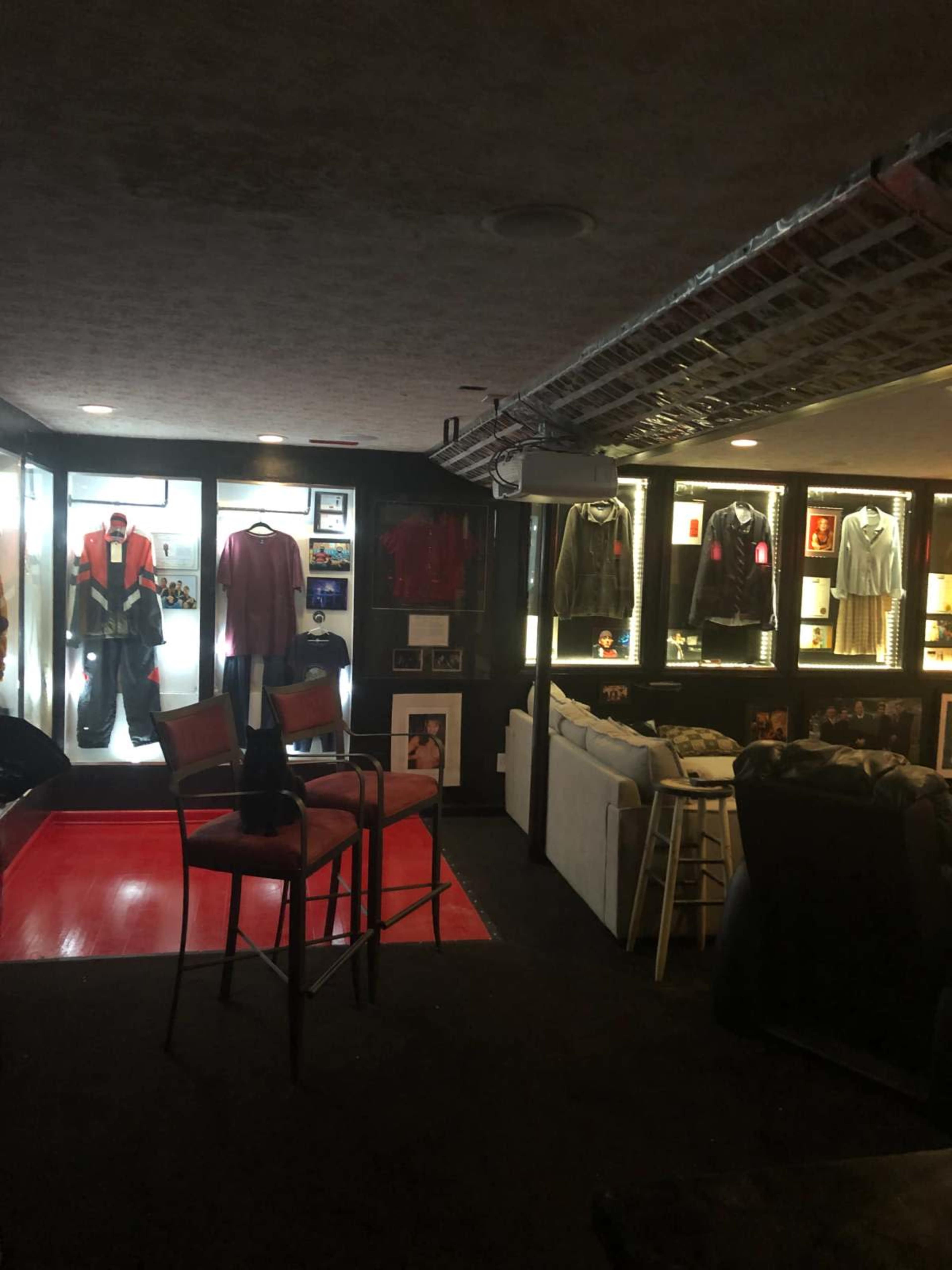 The image shows a dimly lit room featuring display cases with clothing and artwork, along with seating arrangements and a barstool.