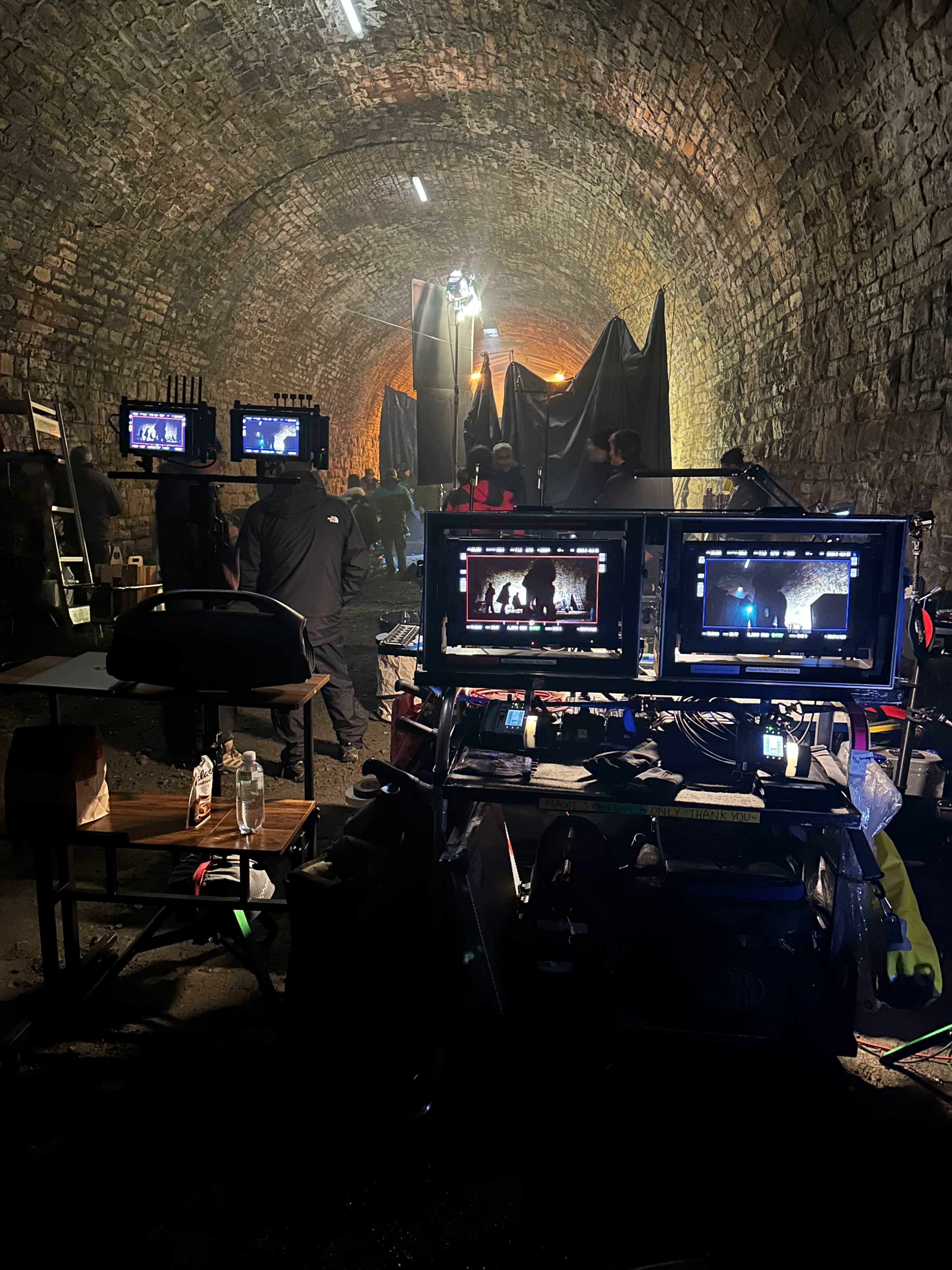 A film set in a dimly lit tunnel, featuring multiple monitors on a table, crew members working, and equipment staged amidst a stone archway background.