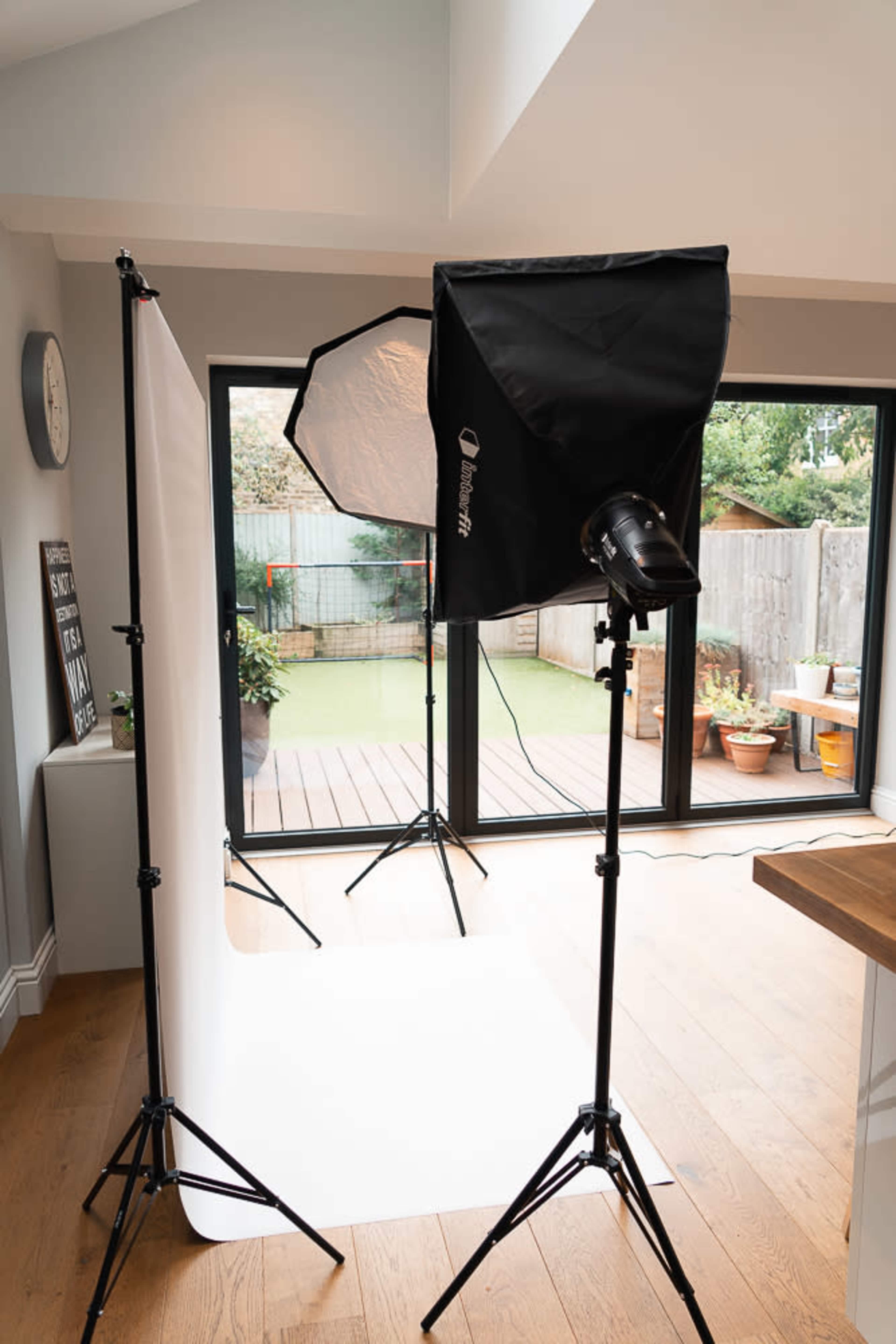 A photography setup in a well-lit room featuring two lights on stands and a white backdrop positioned in front of a large window with a garden view.